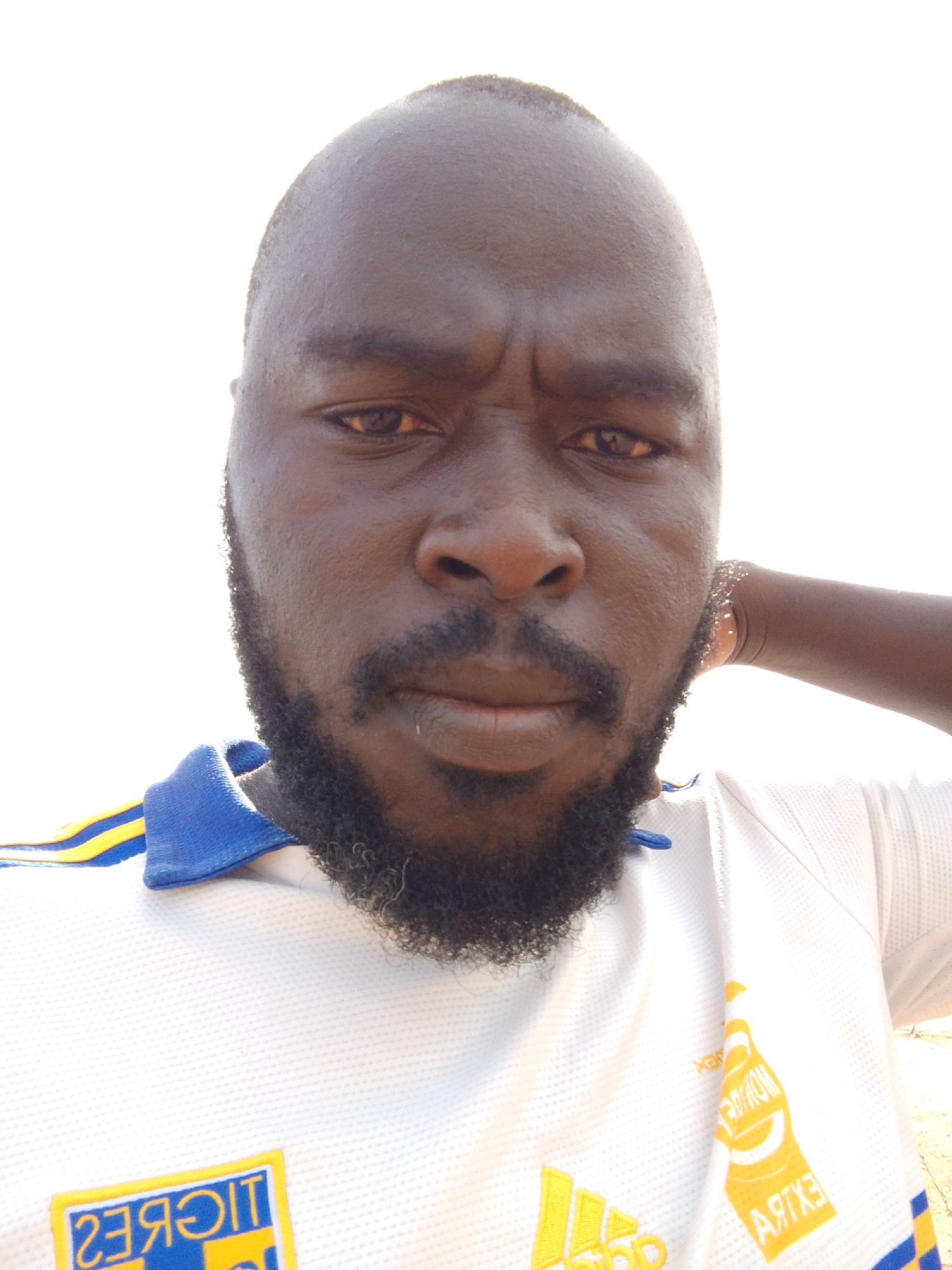 The image shows a close-up selfie of a dark-skinned man with a beard and a furrowed brow. He is wearing a white Adidas t-shirt with blue and yellow accents. The shirt has a Tigres logo and a "Modelo Extra" beer logo. The background is a bright, overexposed white, suggesting strong sunlight. There is no discernible contextual information indicating the exact location within Mulke Barikin Sayejo, Nigeria.