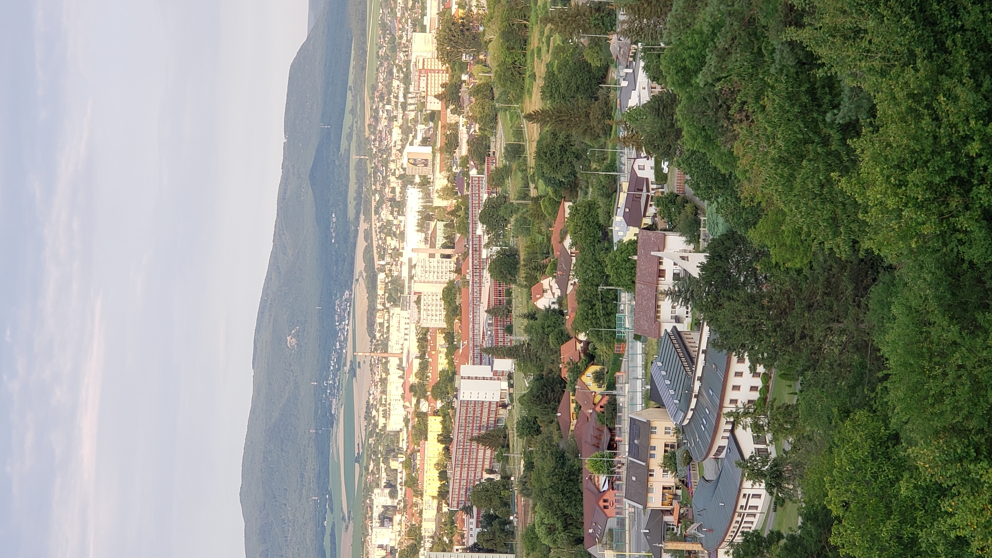 This is an aerial, wide-angle, medium shot taken from a high vantage point overlooking a city. The image shows a layered landscape with foreground trees, a middle ground of residential buildings and a large, long, brick building with many windows, and a background of a city with taller buildings and a mountain range beyond. The sky is a pale blue with some wispy clouds. The overall impression is of a suburban or semi-urban area bordering a larger city and natural landscape. The sun appears to be low in the sky, casting a warm light on the buildings in the distance.
Specifically, this view is from the northeast side of Prešov, Slovakia, looking southwest. The prominent red brick building in the middle ground is the University Hospital in Prešov. The large, curved building in the lower middle ground appears to be part of a residential or institutional complex. The residential houses in the foreground are typical of a suburban neighborhood. The city in the background suggests the main urban center of Prešov. The mountain range in the far background is likely the Slanské vrchy (Slanské Hills).