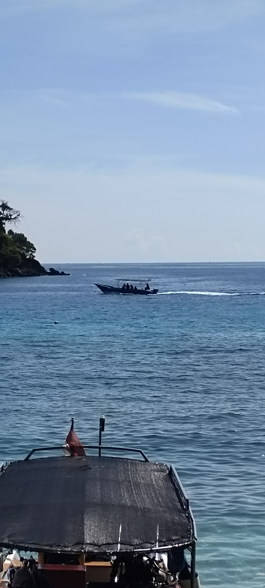 The image depicts a serene seascape off the coast of Sabang, Indonesia, under a clear blue sky. In the mid-ground, a speed boat navigates the tranquil blue waters, leaving a white wake. The boat appears to be carrying several passengers, their silhouettes visible against the open sea. To the left, a lush green island with a rocky shoreline juts into the water, its trees creating a dark, verdant contrast. In the foreground, the partial view of another boat, likely docked or anchored, shows its dark canopy and some equipment, including a red flag. The overall scene evokes a sense of peaceful maritime activity in a tropical setting.