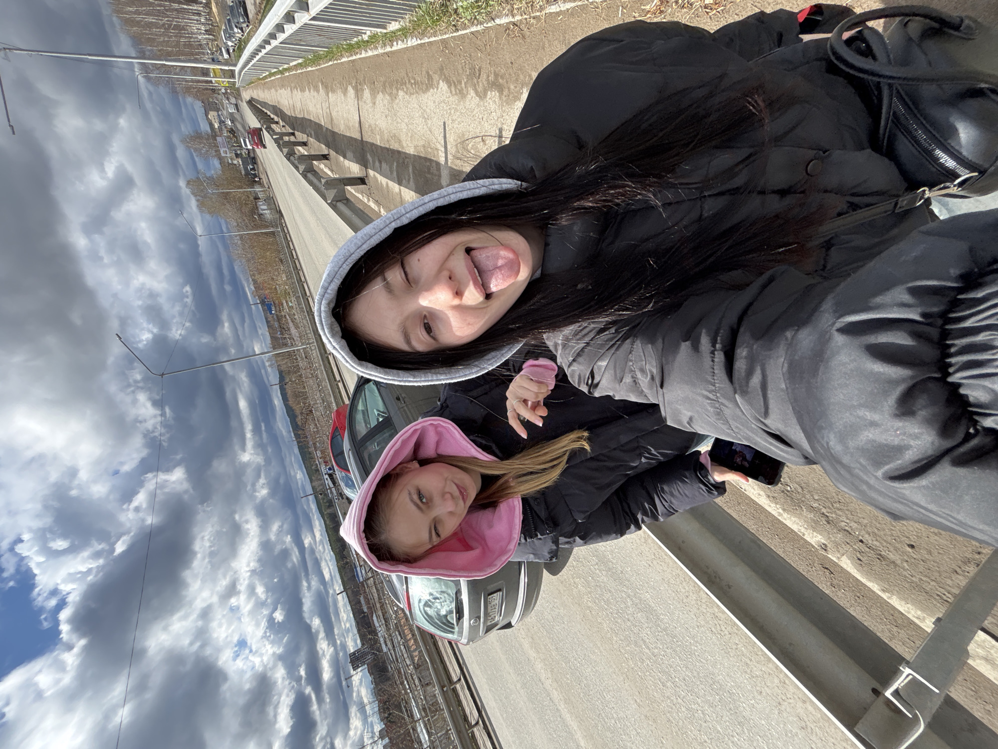 The image displays a playful outdoor scene featuring two young women posing for a photo. The camera perspective is a close-up selfie, taken from a slightly high angle and rotated 90 degrees clockwise, making the subjects appear as if they are standing vertically in a landscape orientation.
In the foreground, a young woman with long dark hair and a black puffer jacket over a grey hooded sweatshirt winks and sticks out her tongue playfully towards the camera. Behind her, slightly to the right, another young woman with lighter brown hair, also wearing a black puffer jacket but with a pink hooded sweatshirt, looks towards the camera with a subtle smile. She is holding a smartphone in her hand.
They are positioned beside a paved road, which features metal guardrails along its edge. A dark-colored car, possibly a hatchback or small SUV, is parked behind the second woman, and part of a red car is visible further back. The background shows an expansive sky with a mix of bright white cumulus clouds and patches of blue. Bare deciduous trees line the distant landscape, suggesting a late winter or early spring setting. Street lights or utility poles are visible along the roadside. The bright sunlight casts distinct shadows from the guardrails onto the ground.
A partially visible license plate on the dark car displays alphanumeric characters, including "602" and "52" (likely a region code), consistent with a Russian vehicle registration. The image's context is Chaikovsky, Russia. The overall mood is casual and cheerful.