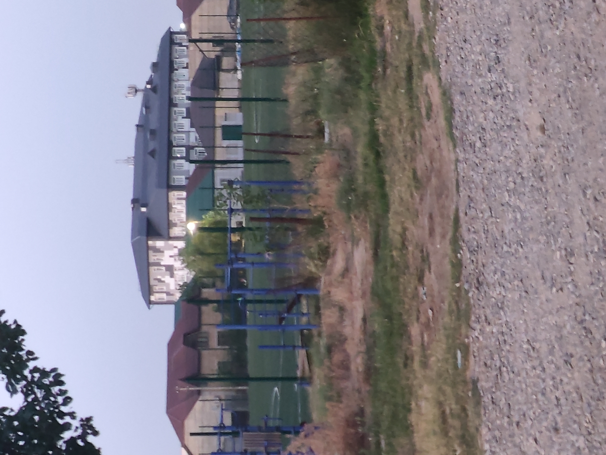 The image captures a park area in Leninkent, Russia, at dusk or dawn. A large, multi-story building with a dark roof and numerous windows dominates the background. In the mid-ground, there is a green sports field with white markings, enclosed by a fence. Adjacent to the field, a playground with blue metal bars and structures is visible. The foreground consists of a gravel path with scattered grass and dry vegetation. A tree with dark leaves is partially visible in the upper left corner. The overall lighting suggests low natural light conditions. There are no people or animals clearly identifiable in the image.