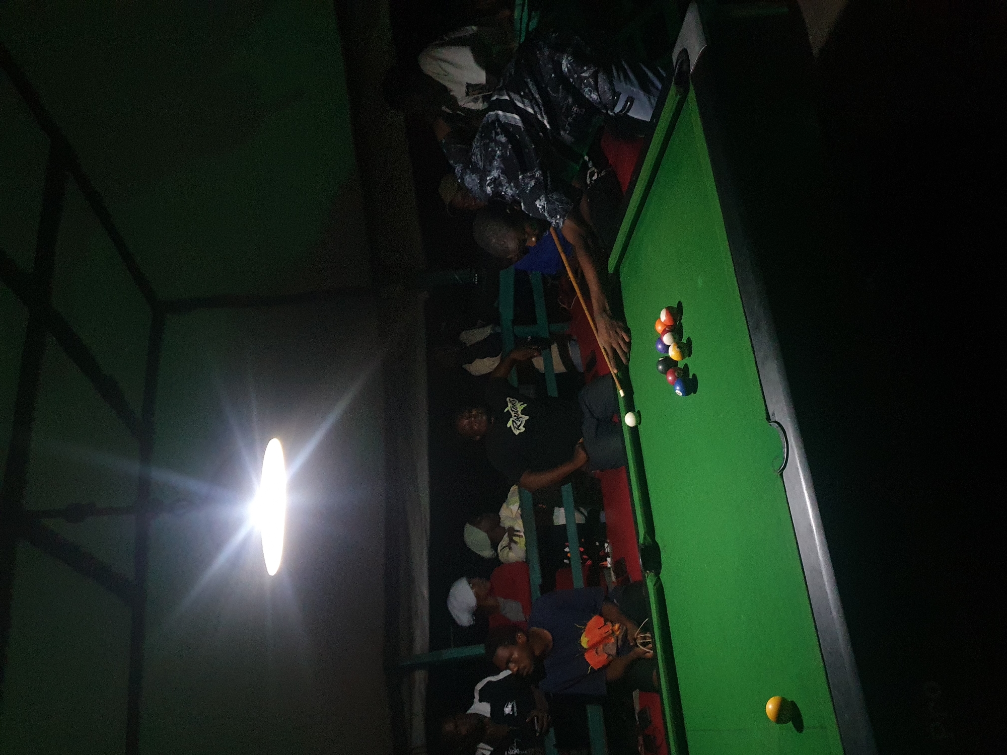 This is a medium shot, outdoor photograph taken at night, likely in Lokoja, Nigeria, depicting a group of people gathered around a pool table under a dimly lit canopy. A bright, diffused light fixture hangs from the ceiling, casting a strong glare.
In the foreground, a green pool table is the central focus. Several billiard balls are scattered across the felt surface, including a yellow cue ball, a red and white striped ball, a blue and white striped ball, a solid red ball, and a solid yellow ball. A man, wearing a dark patterned shirt and light-colored pants, is actively engaged in playing pool. He is bent over the table, holding a pool cue with his right hand and resting his left hand on the table for balance. His gaze is directed at the balls on the table.
In the background, several other individuals are visible, seated and standing around the pool table. They appear to be watching the game. On the left side of the image, a man in a dark blue t-shirt with an orange graphic is seated, looking towards the table. Further back and to the left, a man in a black and white jacket is standing. Several women are seated behind the pool table, some wearing headscarves. Their faces are partially obscured by shadow and distance. The environment appears to be an open-air structure with a dark roof and visible support beams. The overall ambiance is casual and social.