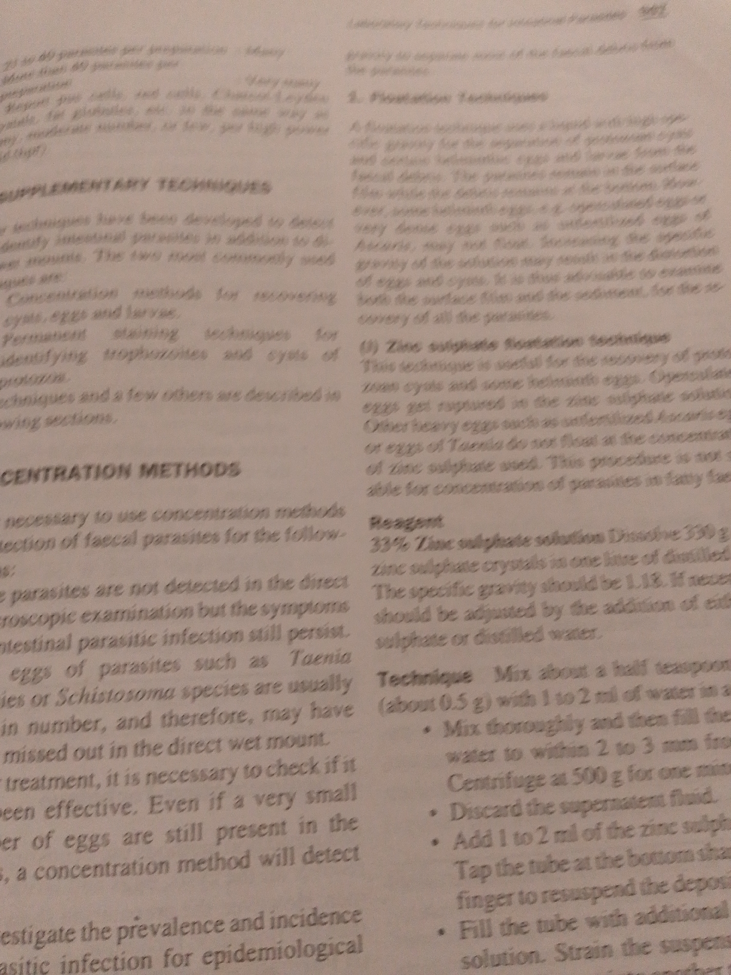 The image is a close-up of a book page with text printed on it. The text is in English and discusses techniques for identifying parasites in fecal samples. There are no people or objects visible besides the book page itself. The scene is a plain, document-like setting, likely a laboratory or study area. The lighting is even, suggesting an indoor environment without any specific time of day indicated. There are no weather cues.
The visible text includes headings such as "SUPPLEMENTARY TECHNIQUES," "CONCENTRATION METHODS," and "Reagent." It also details specific methods like "Zinc sulphate flotation technique" and provides instructions for preparation and use. The text appears to be from a scientific or medical publication. There are no discernible emotions conveyed by the image itself.