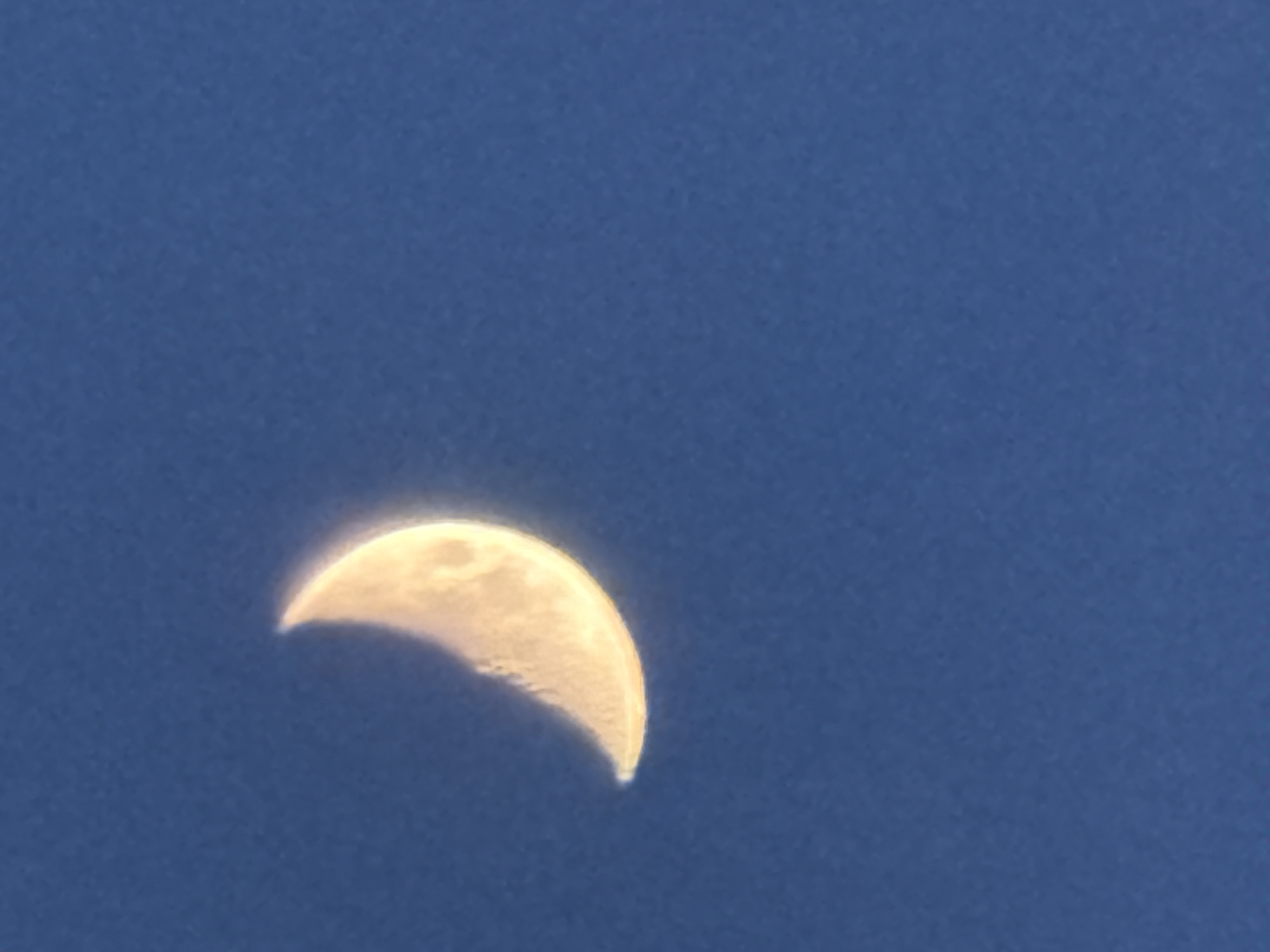 This image features a distinct crescent moon set against a clear, deep blue sky. The moon, positioned in the lower-left quadrant, is illuminated with a bright, yellowish-white light, highlighting its curved form. Visible details on the moon's surface, such as subtle craters and irregularities, are apparent along its illuminated edge, suggesting some texture. A soft, luminous glow emanates from the bright side of the moon, contrasting with the dark curve of the unlit portion. The sky itself is a uniform and cloudless expanse of blue, indicating fair weather. The presence of the moon in a bright blue sky suggests the photograph was taken during daytime, likely in the early morning or late afternoon/early evening hours. There are no other subjects, activities, or visible text in the frame.