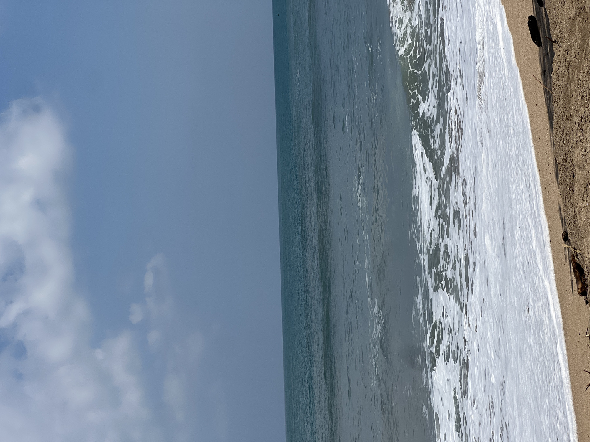 The image shows a tranquil beach scene in Kuta, Indonesia. The calm, turquoise ocean stretches to the horizon, meeting a pale blue sky dotted with wispy white clouds. Gentle waves lap at the sandy shore, creating a foamy white border. The sand is a light brown, with a few pieces of driftwood scattered near the water's edge. No people are visible in the image, adding to the serene atmosphere.