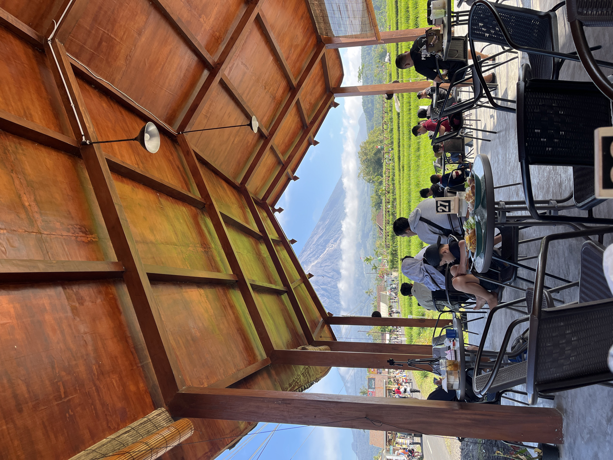 The image depicts an outdoor dining area with a wooden roof supported by wooden beams and pillars. The ceiling is made of polished wood, with visible planks and some areas showing signs of weathering or moss. Hanging pendant lights are suspended from the ceiling. In the background, a large, majestic mountain, likely Mount Merapi, dominates the skyline, with its peak partially obscured by clouds.

The foreground is populated by several tables and chairs, mostly made of dark-colored woven plastic or metal. People are seated at some of the tables, engaged in eating, drinking, or conversing. A young boy is visible sitting with his legs crossed, looking down at a plate of food. Another man is standing near a table, facing away from the camera.

Beyond the dining area, lush green rice fields stretch across the landscape, indicating a rural or semi-rural setting. A road runs alongside the rice fields, with several vehicles and pedestrians visible, suggesting a pathway or a small town. The overall atmosphere appears relaxed and casual, with people enjoying a meal or a break amidst a scenic natural environment. The location is identified as Oro-oro Ombo, Indonesia, a village known for its proximity to Mount Merapi.