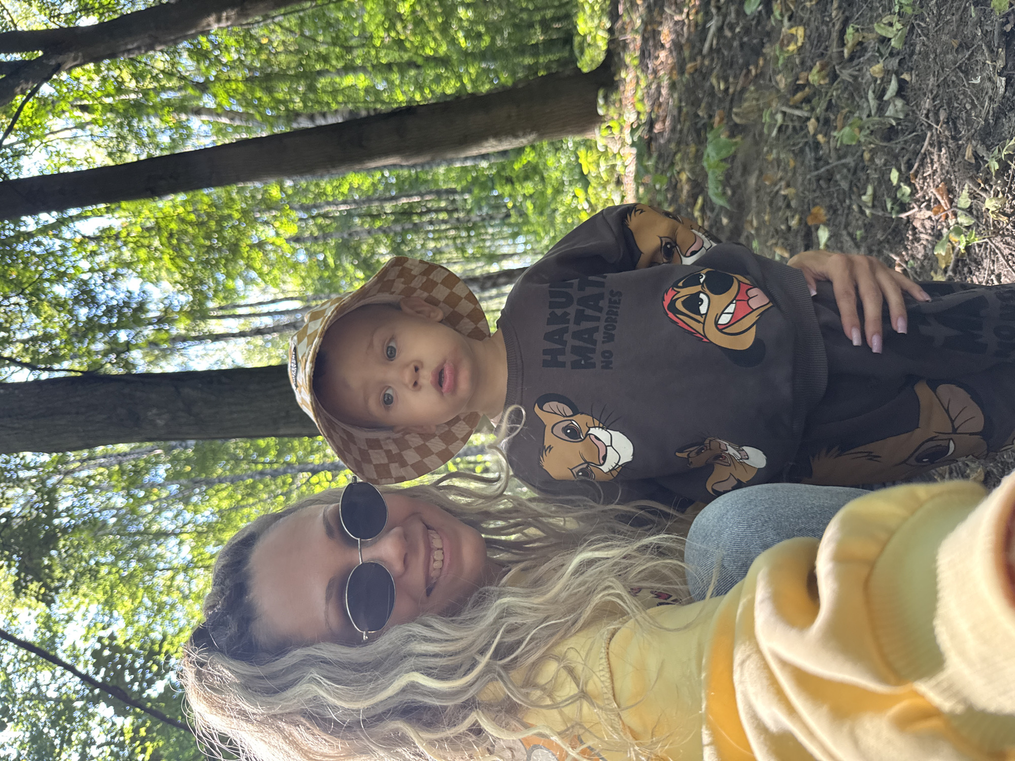 In Rusanovo, Russia, a woman with blonde curly hair and sunglasses is taking a selfie with a young child. She is wearing a yellow hoodie, and the child is wearing a patterned bucket hat and a dark brown sweatshirt with characters from "The Lion King" printed on it. They are in a forest with tall trees and dappled sunlight filtering through the leaves. The ground is covered with dry leaves and twigs. The woman has her arm around the child, and they are both looking towards the camera.