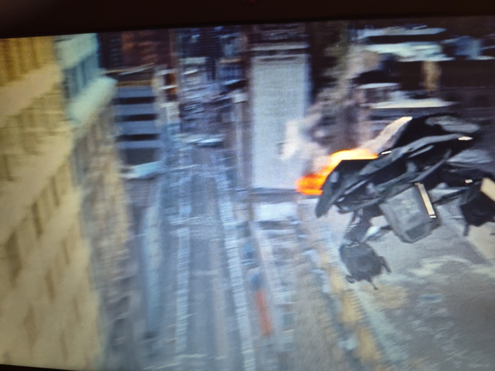 The image shows a blurry aerial view of a city with tall buildings. In the foreground, a dark, angular object, possibly a vehicle or aircraft, is visible with some orange flames or lights emanating from it. The overall impression is one of an urban environment with some kind of action or event taking place. There is no visual information in the image that can identify the location as Sandwell, United Kingdom.