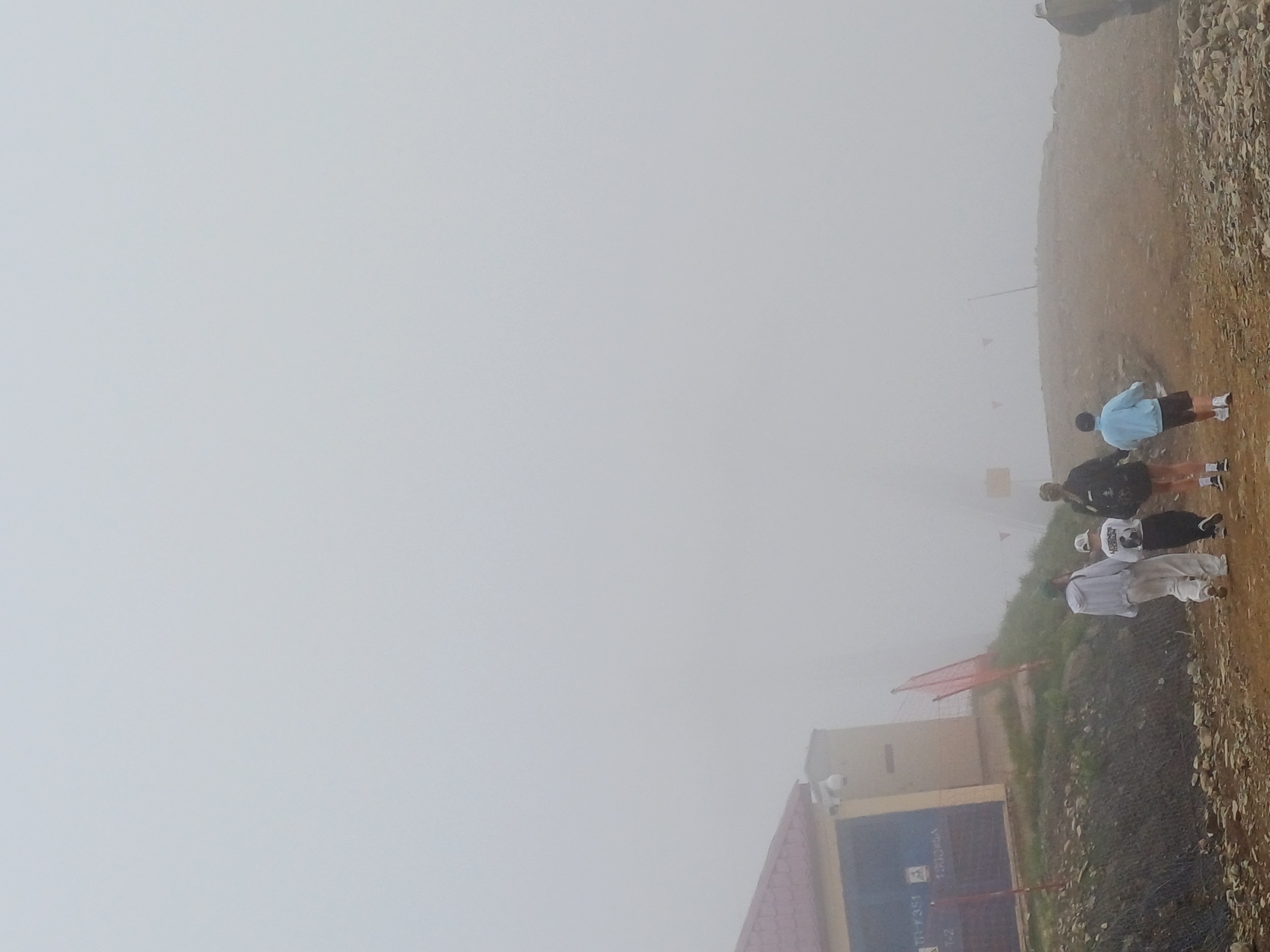 The image captures a group of people on a rocky, outdoor slope, partially obscured by fog. Four individuals are clearly visible, with at least one more person in the background. One person, wearing a light blue jacket and dark shorts, stands facing away from the camera. Another person in a white outfit and a white hat is to their left, also facing away. Further back, two more figures are visible, one in black and another in white. The setting appears to be a mountainside or elevated terrain, with loose rocks and sparse vegetation. In the background, a building with a blue facade and a red-roofed structure are partially visible through the mist. A metal fence runs along the slope in the foreground. The weather is foggy, creating a muted and hazy atmosphere. There is some visible text on the blue facade of the building, but it is not legible. The overall mood is subdued due to the weather conditions.