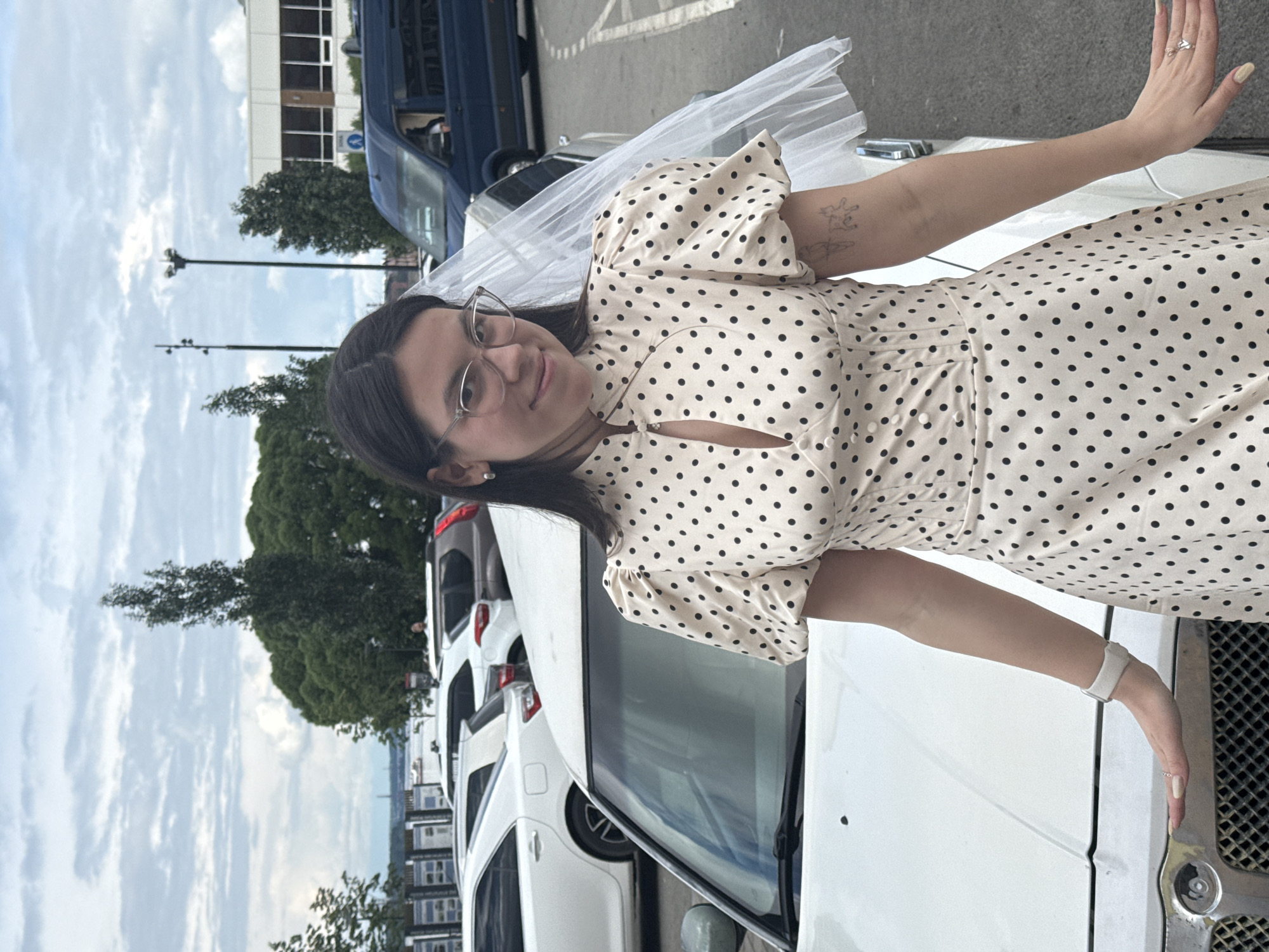 A young woman is prominently featured, positioned centrally and leaning on the front grille and hood of a white Chrysler 300C car. She has dark hair, wears clear-rimmed glasses, and a beige polka-dot dress with puffed sleeves and a high collar with a keyhole cutout. A white veil is draped over her left shoulder, and she exhibits a slight smile towards the viewer. A light-colored tattoo is visible on her upper left arm. She wears a white wristband or watch on her left wrist and rings on her fingers.
The setting is an outdoor parking lot under an overcast sky. Behind the primary subject and her vehicle, several other cars are parked, including a white sedan (possibly a Toyota Camry), a white SUV, a dark brown SUV, and a dark blue van. In the mid-ground, a dense line of green trees separates the parking area from what appears to be a body of water, likely the Kama River, discernible in the distance. A faint structure, possibly a bridge or industrial facility, is visible on the far horizon over the water. To the right, a light-colored building with large windows and a blue circular sign containing a white letter "A" (indicative of a public transport stop or related signage common in Russia) is partially visible. The overall environment, including the architecture, vehicles, and riverfront, is consistent with an urban location in Perm, Russia.