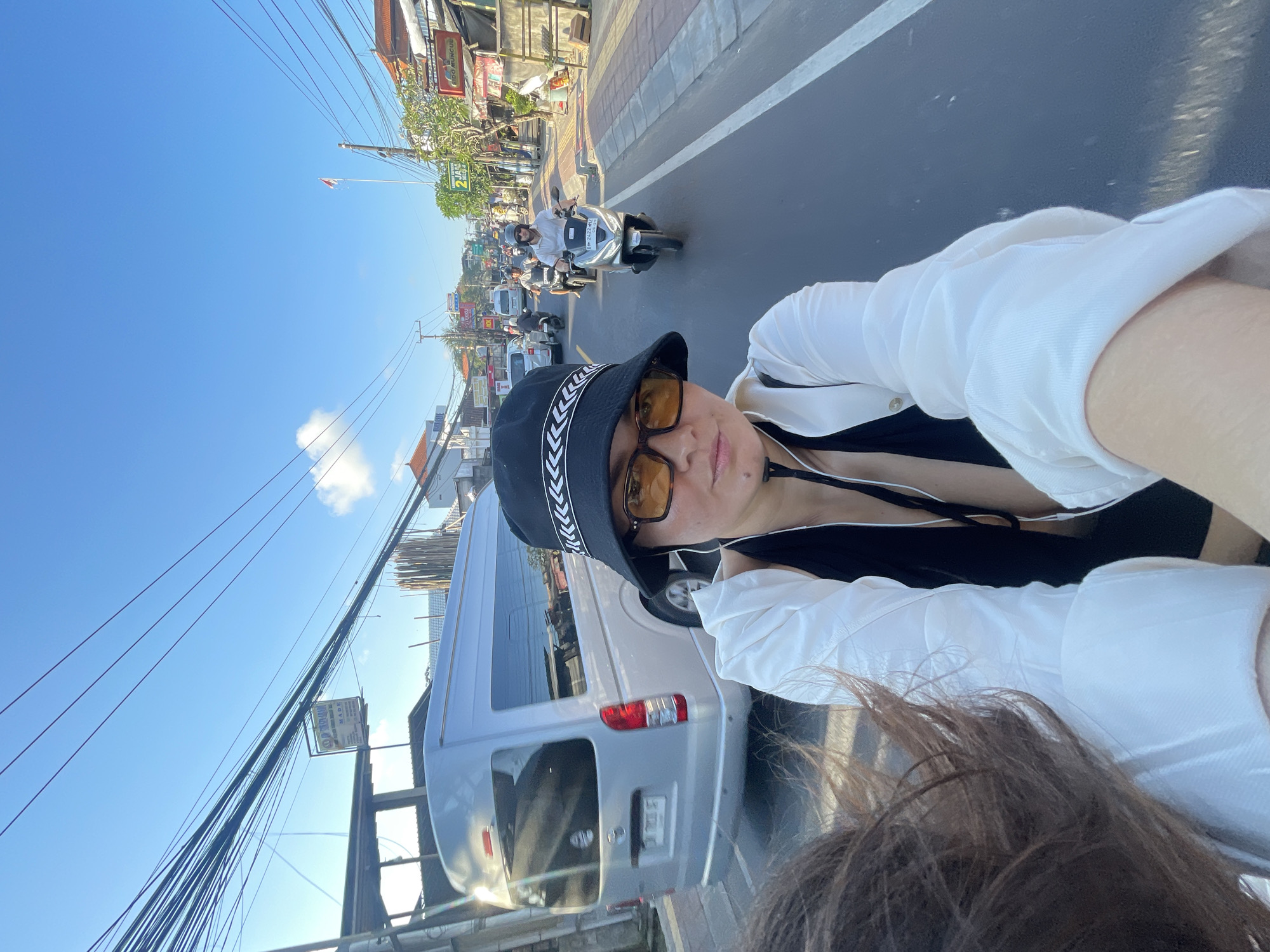 The image is a selfie taken from the perspective of someone riding on the back of a motorbike, with a second person visible in the foreground on the left. The person in the foreground, facing the camera, is wearing a black bucket hat with a white stripe pattern, large orange-tinted glasses, and a white open shirt over a black top. They are in a busy street in Kuta Selatan, Indonesia, with several motorbikes and a silver minivan visible in the background. The street is lined with buildings and shops, and there are numerous overhead power lines. The sky is blue with a few clouds, indicating it is daytime. The overall scene depicts a typical street view in a bustling urban area of Indonesia.