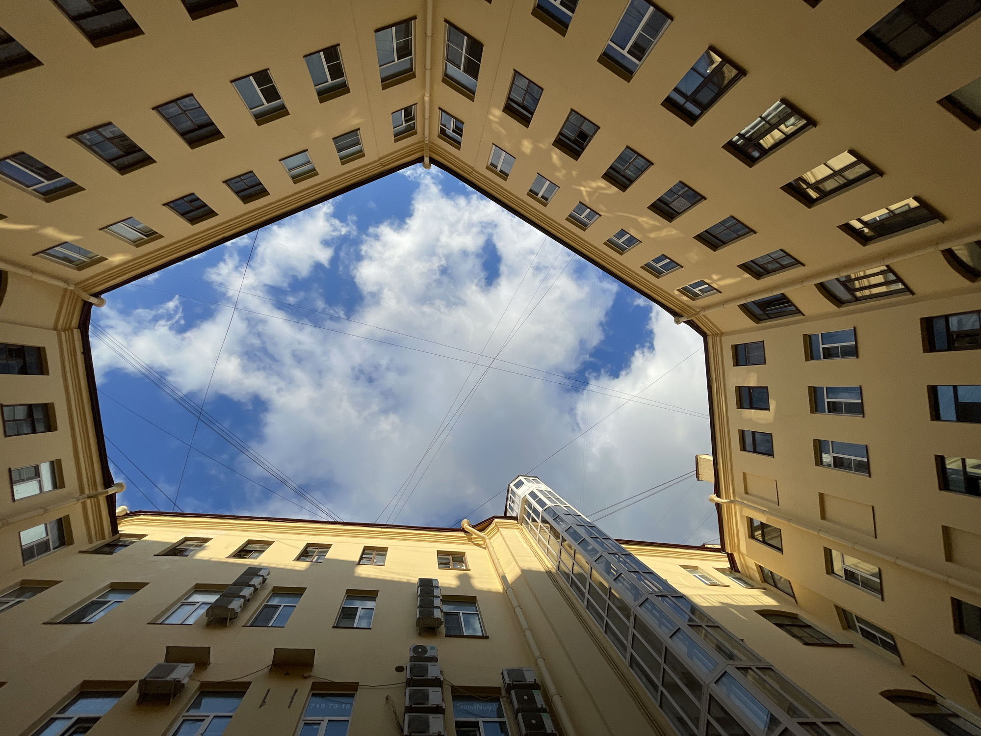 The image depicts a view looking upwards from a courtyard in Saint Petersburg, Russia. The architecture is characteristic of the city, with tall, pale yellow buildings forming a courtyard. The buildings have multiple floors with numerous windows, many of which appear to be of a traditional style with dark frames. The sky above is blue with scattered white clouds, and power lines are visible crisscrossing the sky. An external glass elevator is attached to one of the buildings, adding a modern element. Several air conditioning units are mounted on the exterior walls of the buildings. The perspective is from ground level or slightly below, emphasizing the height and enclosure of the courtyard.