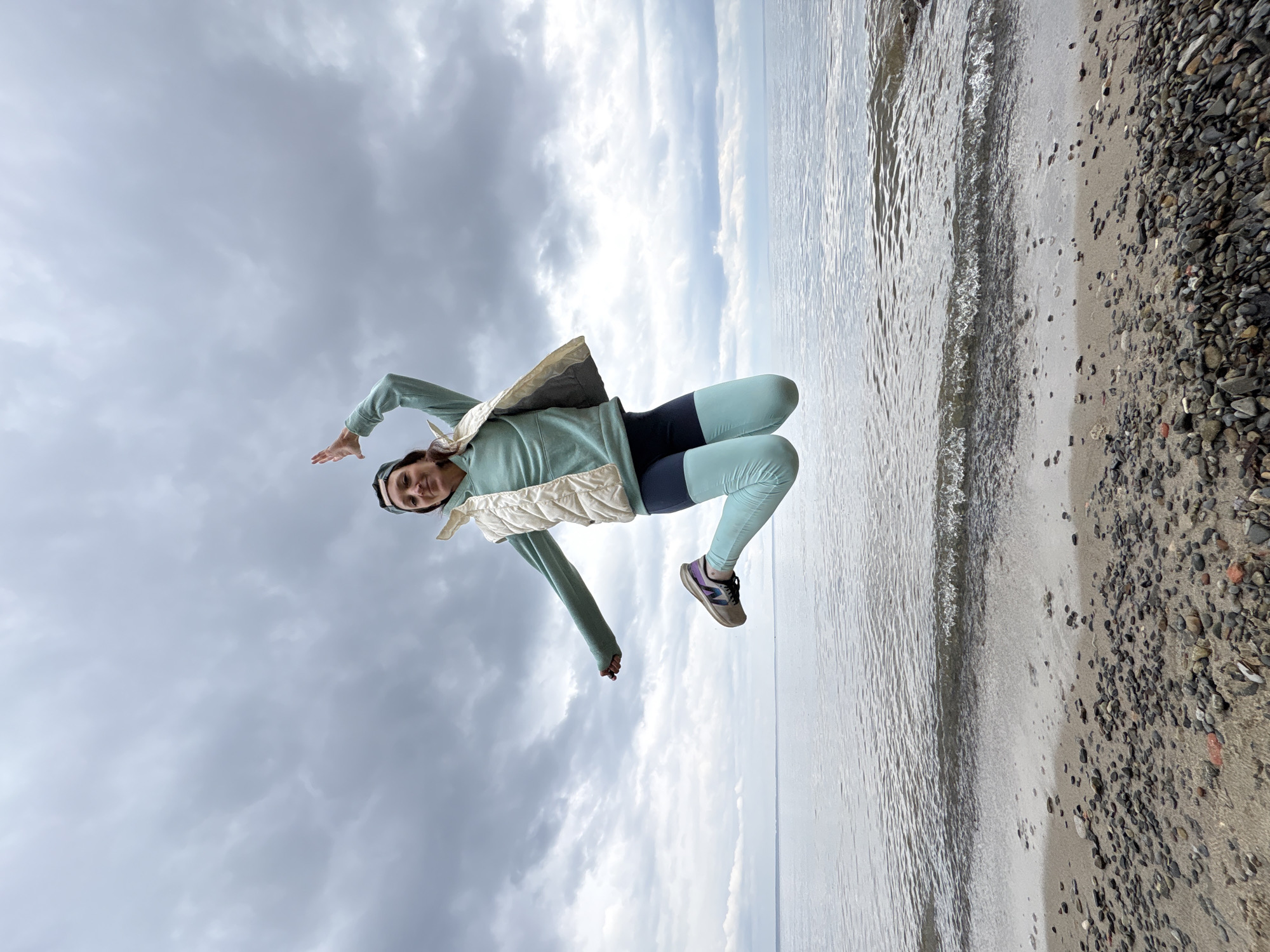 A woman is jumping on a beach near water. She is wearing a light green jacket, a white vest, dark leggings, and sneakers. The beach has a mix of sand and pebbles. The sky is overcast with dark and light clouds. The location appears to be a beach with a body of water, possibly the Volga River, which runs through Ulyanovsk, Russia. However, the precise location within Ulyanovsk cannot be determined solely from the image.