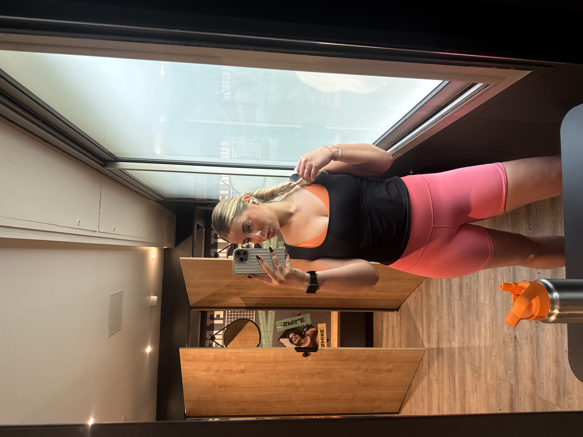 A blonde woman stands in a gym in Paris, France, taking a selfie in a mirror. She is wearing a black tank top over an orange sports bra, pink shorts, and an Apple Watch on her left wrist. She holds a silver iPhone in her left hand. Her blonde hair is pulled back in a braid. In the background are wooden shelves and a water bottle. The gym has a bright window above the shelves.