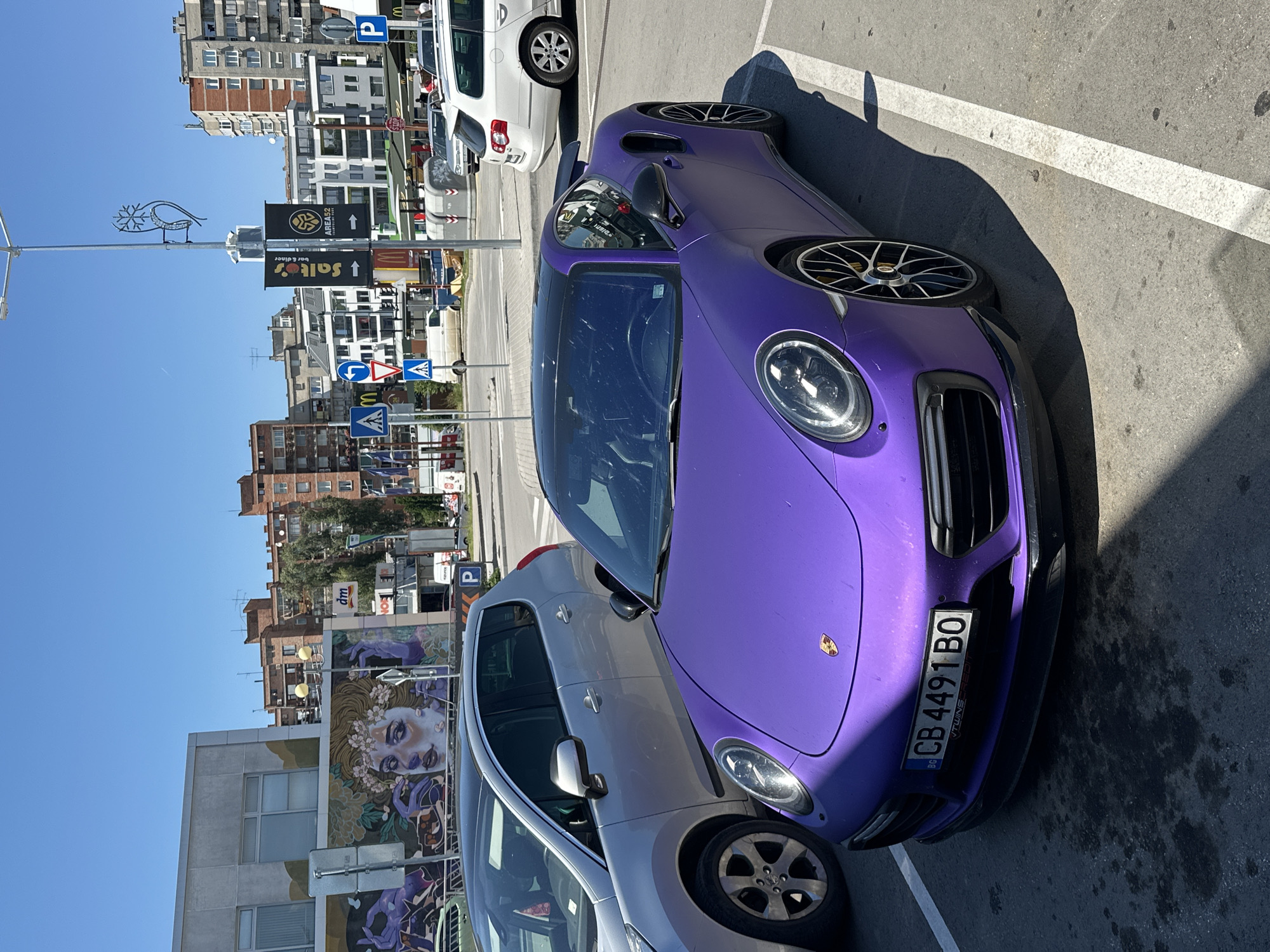 A matte purple Porsche 911 Turbo S is parked in a lot on a sunny day. The car has a black roof and black wheels with yellow brake calipers. The license plate reads "CB 4491 BO". To the left of the Porsche is a silver hatchback car. In the background, there are several buildings, including apartment blocks and commercial establishments. A large mural depicting a woman's face and hands is visible on the side of one building. Signs for "dm", "Salt's Bar & Diner", and "AREA52" are also present. The scene appears to be a street or parking lot in the city of Veliko Tarnovo, Bulgaria.