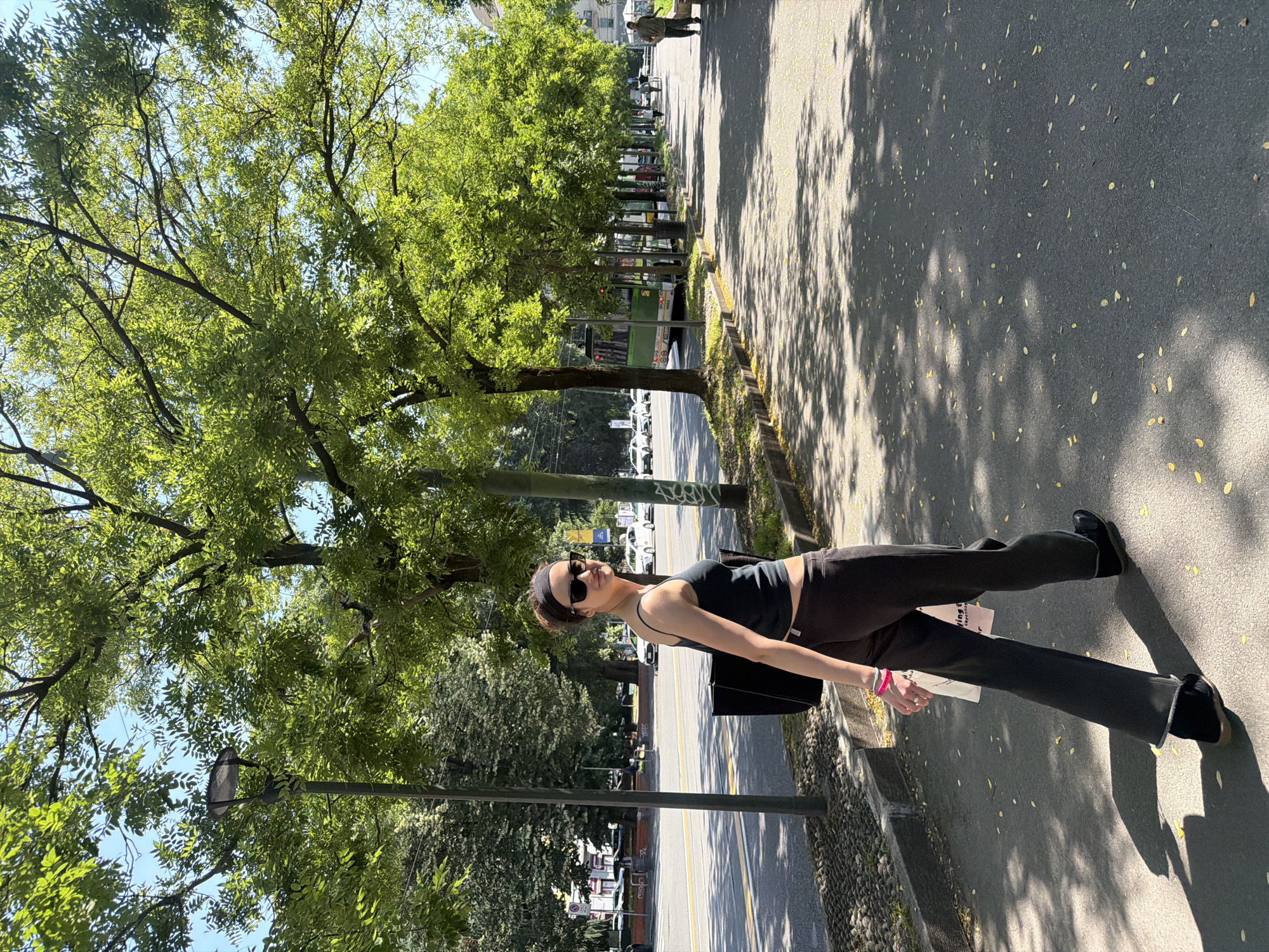 A woman stands on a sidewalk next to a street lined with trees. She wears a black tank top, dark pants, and black shoes. She has sunglasses on and is holding a white object in her left hand. The street has yellow lines and trees line one side of the street. Cars are visible parked along the street. Shadows from the trees are cast on the sidewalk. The location appears to be a street in Milan, Italy.