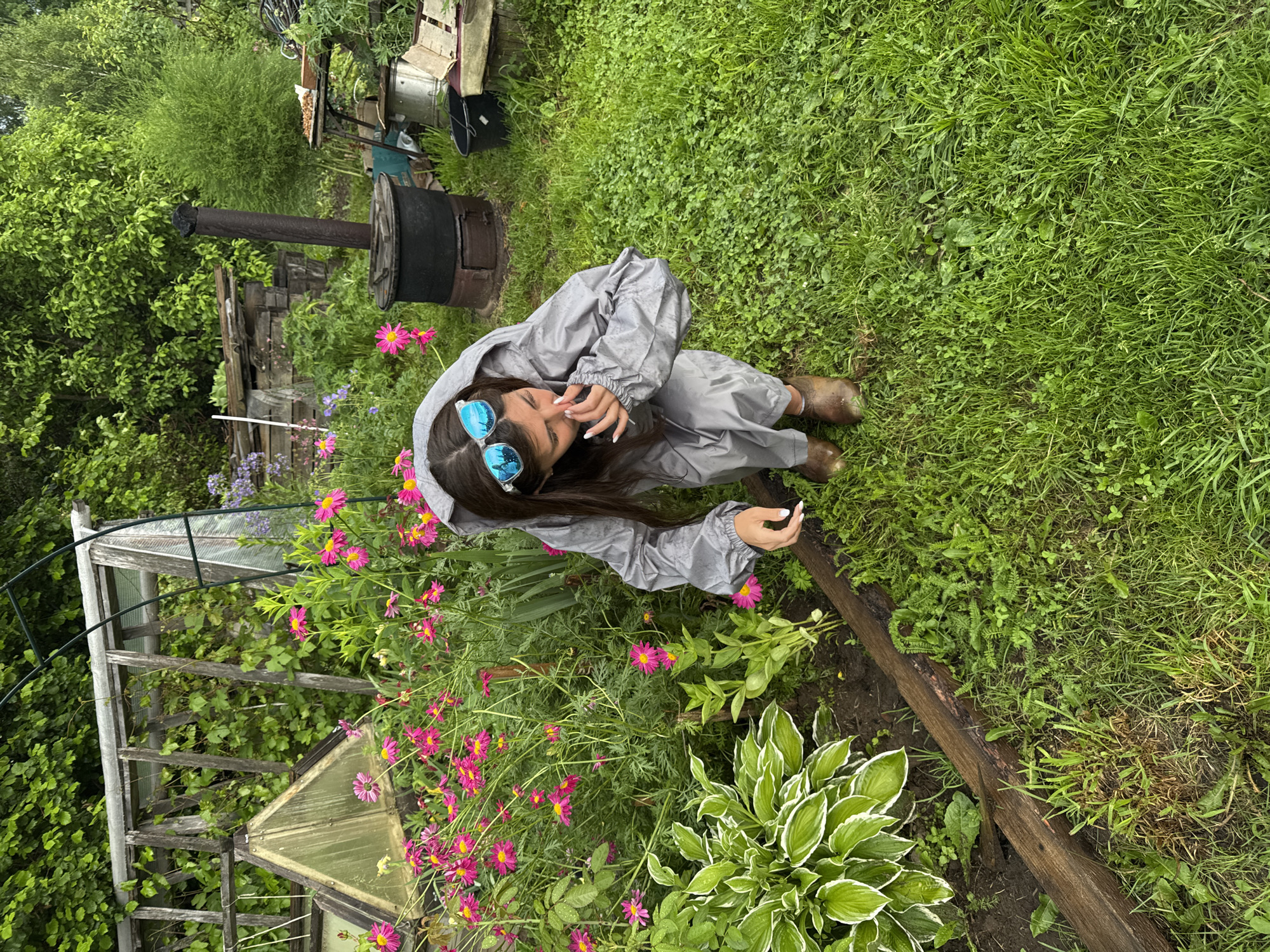 A young woman wearing a gray hooded rain suit and brown boots is crouched in a garden in Вассята, Russia. She is wearing large blue reflective sunglasses and has her hand near her nose. She is surrounded by lush green grass, pink flowers, and various garden structures including a greenhouse and a black metal stove. The environment appears to be a backyard or rural garden setting.
