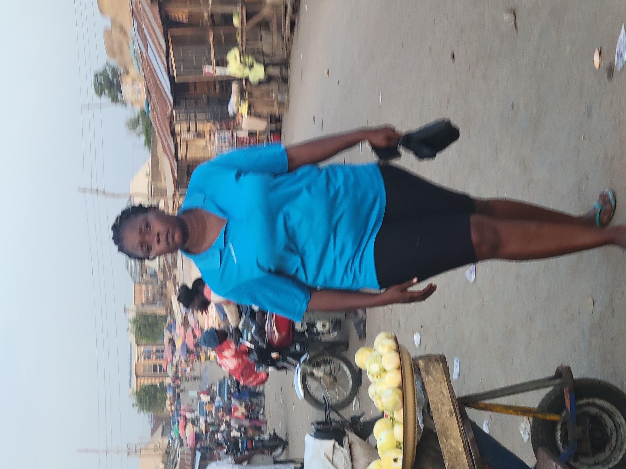 An outdoor, daylight shot depicts a woman in Kwamba, Nigeria, standing near her merchandise on a busy street. The woman, wearing a blue shirt and black skirt, stands next to a handcart filled with yellow fruits, possibly mangoes or pears, ready for sale. Behind her, a red motorcycle is parked amidst the pedestrian traffic. The street scene is filled with people and various stalls, indicating a bustling marketplace. In the background, buildings and power lines are visible under a hazy sky.