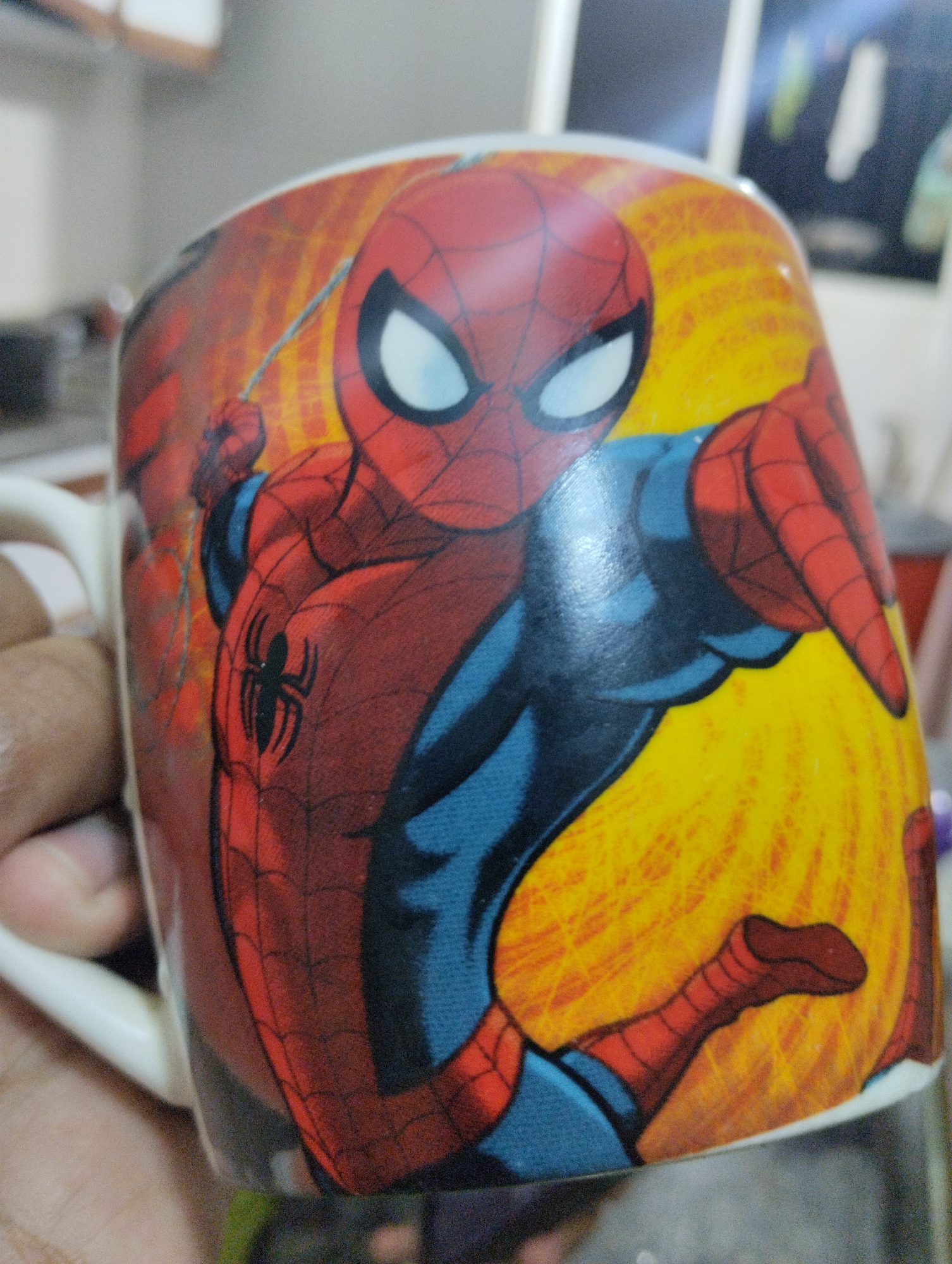 The media file primarily features a white ceramic mug adorned with a vibrant depiction of the Marvel superhero Spider-Man. A human hand, with a light brown skin tone, is shown holding the mug; the thumb and part of the palm are visible on the left side.
The artwork on the mug presents Spider-Man in his iconic red and blue suit. His mask is red with large white eye lenses outlined in black. The character's upper body and limbs are predominantly red with black web-like patterns, while the midsection, parts of the arms, and legs are solid blue. A prominent black spider emblem is clearly visible on his left chest. Spider-Man is depicted in a dynamic, action-oriented pose, with his left arm extended forward, and a thin white line, suggesting a web, emanates from his wrist. The background design on the mug is a bright, radiating yellow-orange, conveying energy.
The setting is indoors, indicated by the heavily blurred and indistinct background, which suggests a room environment with subtle hints of furniture or walls. No specific location cues directly related to Caracas, Venezuela, are discernible from the visual content. There are no indications of the time of day or weather, nor are any emotions overtly conveyed. No visible text is present on the mug or in the background.