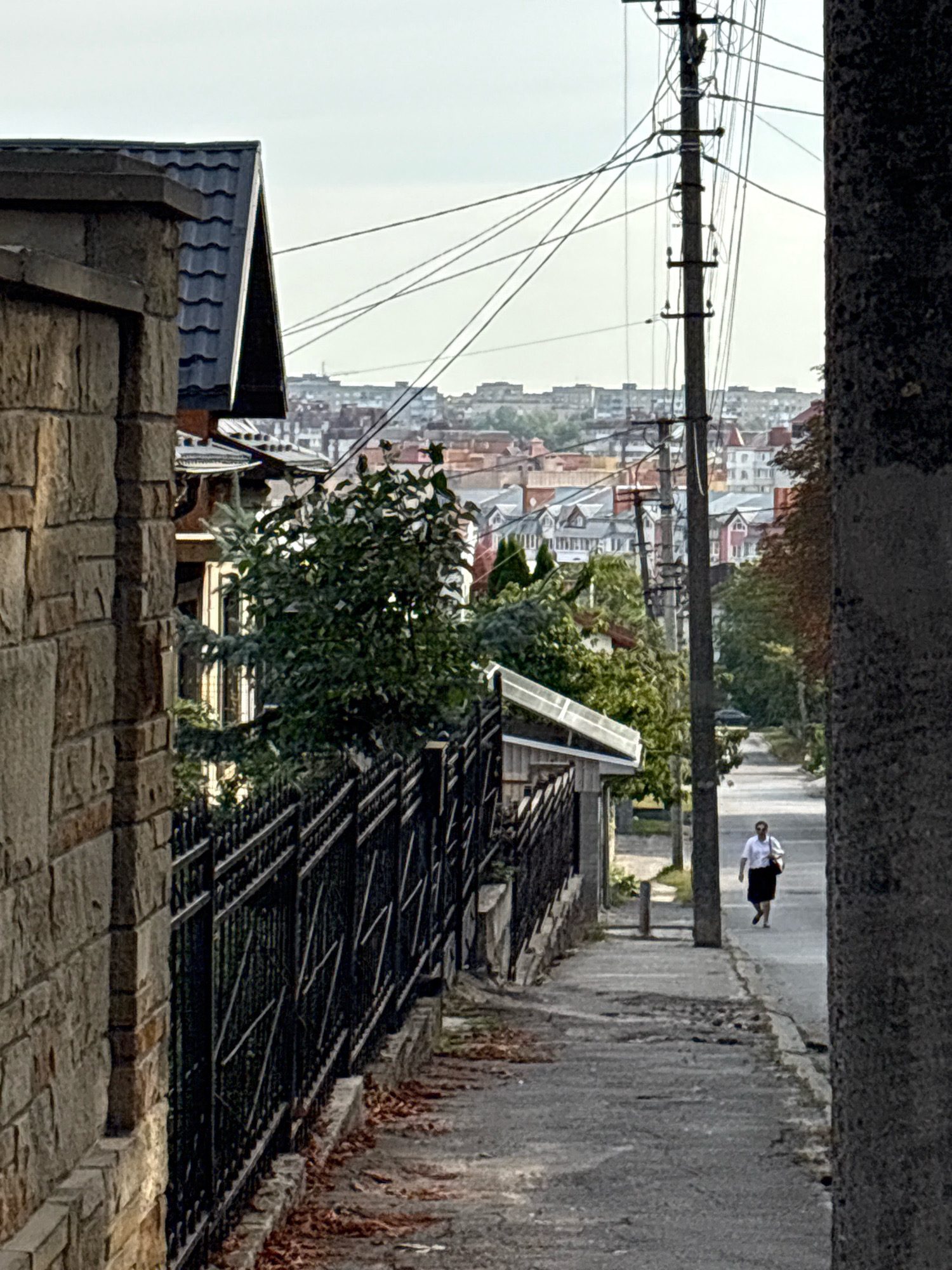 The image depicts a street scene in Ternopil, Ukraine. In the foreground, a stone wall and a black metal fence line the left side of the path. The path itself appears to be a paved sidewalk transitioning into a street. Further down the street, a single person, a woman wearing a white blouse and a dark skirt, is walking away from the camera, carrying a light-colored bag over her shoulder. A wooden utility pole with numerous wires stretches vertically along the right side of the street. In the background, a dense collection of residential buildings with various colored roofs is visible, suggesting a moderately populated urban area. The sky is overcast. The environment appears to be an outdoor, daytime setting.