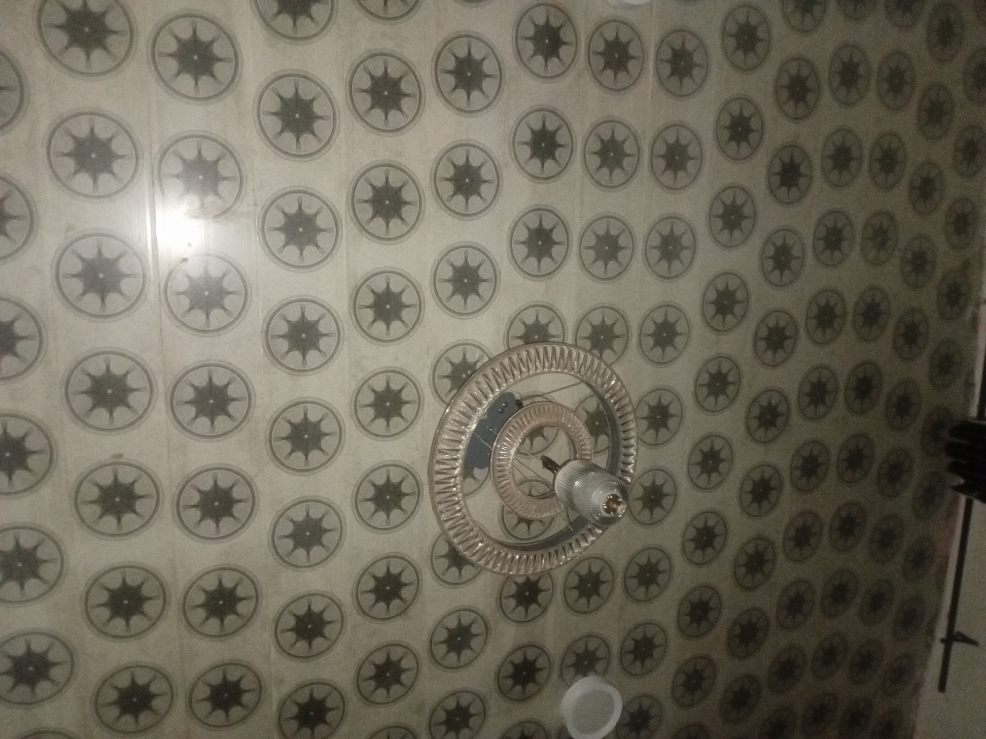 This image captures the interior ceiling of a room in Ogbogbo, Nigeria, viewed from a low-angle perspective looking upwards. The ceiling is uniformly covered with light-toned rectangular panels, likely PVC or similar material, arranged in a grid. Each panel features a dark, repeating circular motif containing a stylized eight-pointed star or sunburst design, set against an off-white or light grey background.
A modern chandelier is suspended centrally from the ceiling. It is constructed from multiple concentric, clear, possibly acrylic or glass, rings, with the outermost ring displaying a ribbed or textured edge. A single, elongated light fixture is mounted at the core of these rings, enclosed in a clear or frosted shade, providing artificial illumination. Reflections from this light source are visible on the surrounding ceiling panels and the polished surfaces of the chandelier. To the left of the chandelier, a partial white, inverted dome-shaped object, possibly a light cover or vent, is visible. The very bottom edge of the image shows indistinct dark architectural elements, likely part of a wall or another fixture below the ceiling line. No individuals are present in the scene.