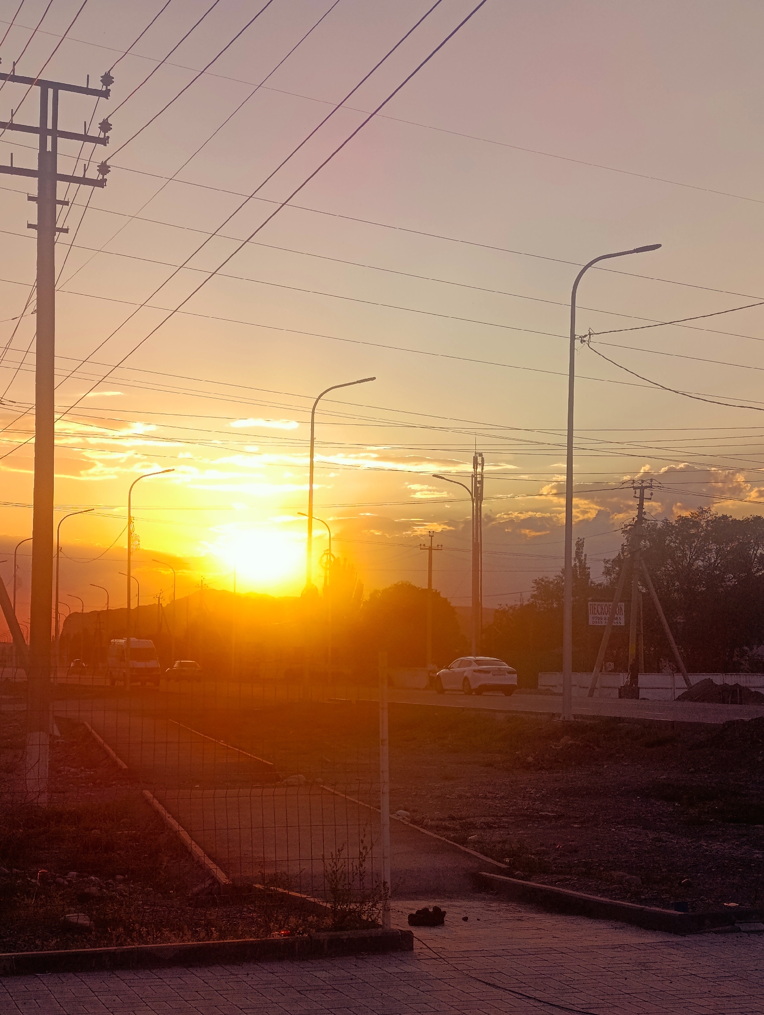 The image depicts a street scene at sunset in Naryn City, Kyrgyzstan. The sun is low on the horizon, casting a warm, golden glow across the sky and illuminating the clouds. Several power poles with wires crisscross the frame, along with streetlights. On the road, a white sedan and a van are visible, suggesting vehicle traffic. The foreground shows a paved walkway or sidewalk and a fenced-off area with gravel and vegetation. In the background, trees and more power structures are silhouetted against the sky. A sign with Russian text is mounted on a pole on the right side of the image. The overall atmosphere is one of a typical urban environment transitioning into evening.