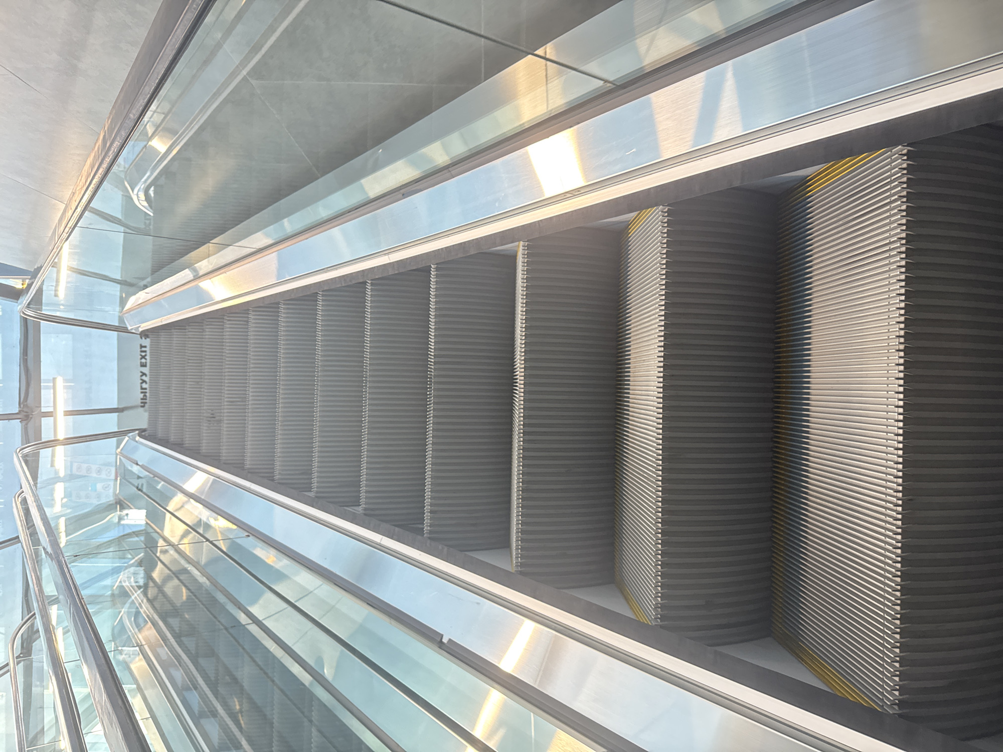 An upward-view shot captures the steps of a modern escalator in Bishkek, Kyrgyzstan. The escalator features a sleek design with polished metal and glass railings, reflecting the bright, natural light filtering from above through a translucent ceiling. The steps themselves are a metallic gray, with yellow safety lines marking the edges. The angle of the shot emphasizes the endless upward progression of the escalator, creating a sense of depth. In the background, barely visible through the glass, is text that appears to read "EXIT." The overall environment suggests a public space, likely a shopping mall, airport, or transportation hub.