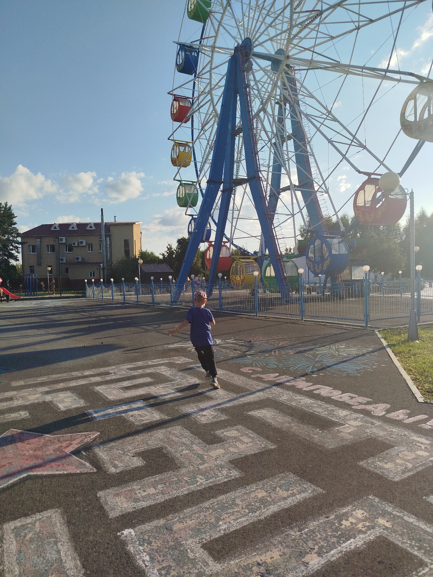 This image shows a child running across a chalk-drawn maze on the asphalt in front of a Ferris wheel. The Ferris wheel is tall and blue, with multi-colored gondolas. Behind the Ferris wheel, there is a building with a red roof and several air conditioning units. The sky is blue with some scattered clouds. The scene appears to be a public park or amusement area. The text on the maze is in Russian, possibly "САМАЯ ПОБЕДА" which translates to "The greatest victory." The location is the city of Yalutorovsk, Russia.