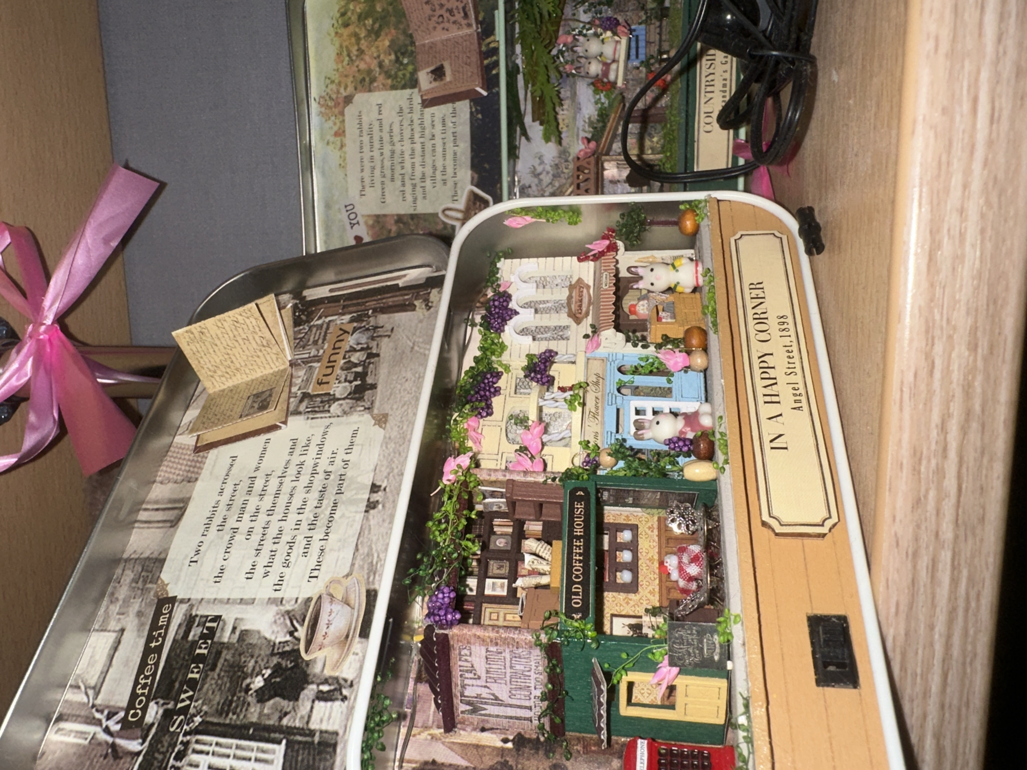 The image shows a diorama inside a tin, likely a craft or display. The diorama features miniature buildings resembling a street scene with shops. One building is labeled "METCALFE BUILDING CONTRACTORS" and another "OLD COFFEE HOUSE." There are tiny figures, including rabbits, and decorative elements like grapes and flowers. A pink ribbon is tied in a bow at the top of the tin. The tin itself appears to be a hinged lid style, with printed images and text on the interior. One printed section has text that reads, "Coffee time SWEET" and a narrative about two rabbits. Another section has text that starts with "YOU" and continues with a story about rabbits. The diorama is situated on a wooden surface, with a second tin visible behind it that also has a printed landscape and text. There is a black electrical cord coiled on the right side of the image. The overall impression is of a whimsical, handmade scene. The location of Petropavlovsk-Kamchatsky, Russia, is not discernible from the visual information provided in the image.