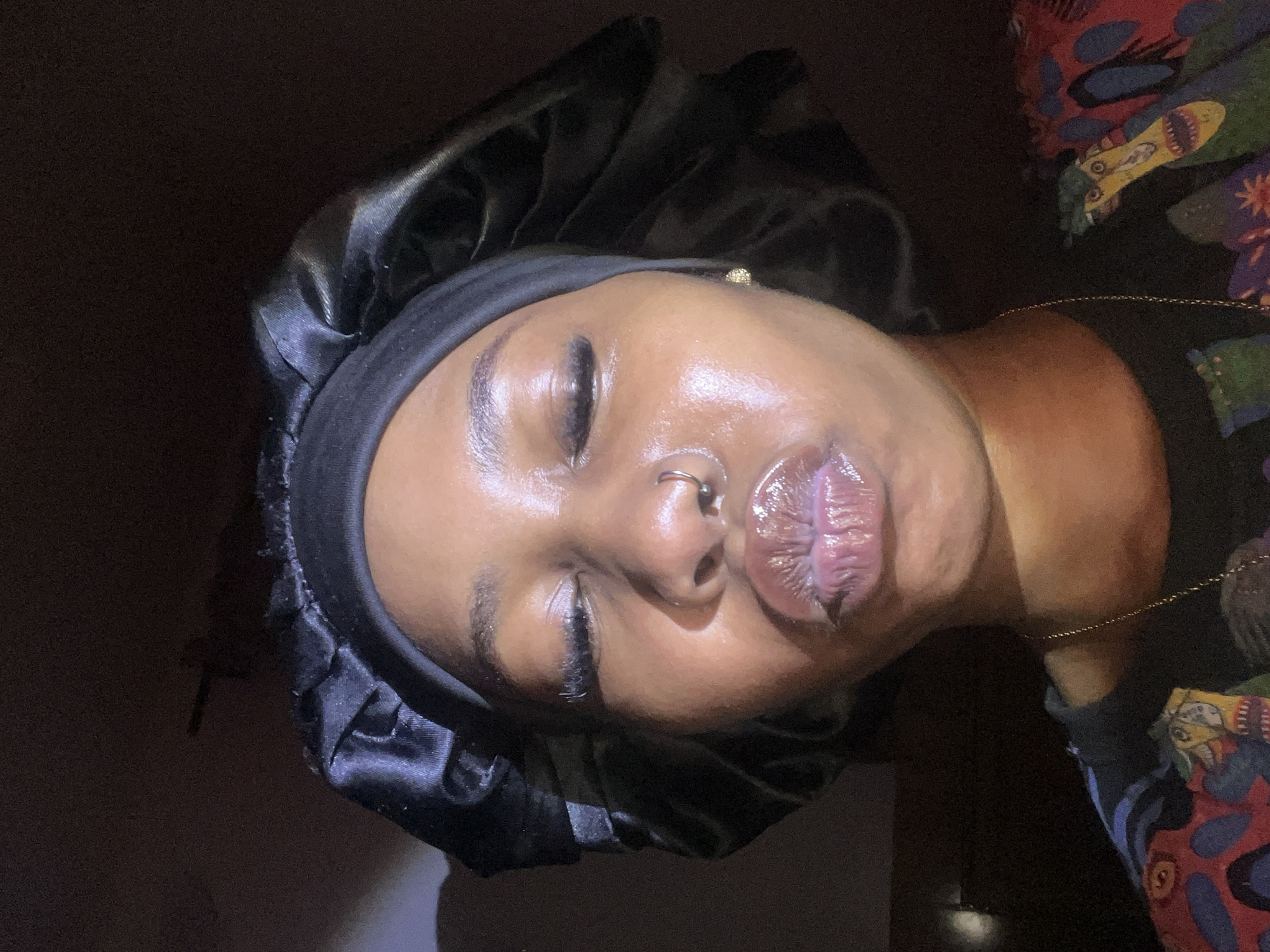 The media file shows a close-up, sideways shot of a person wearing a black satin bonnet. The person's eyes are closed, and they appear to be resting. The lighting is dim, suggesting nighttime or an indoor setting. The person has their lips slightly parted and appears to be wearing lip gloss. A nose ring is visible. On the right side of the image, a colorful, patterned fabric is visible, possibly clothing or bedding. There are no visible text elements in the image. The scene appears to be a personal, intimate moment, possibly before sleep or upon waking. The overall mood is peaceful and relaxed. The location context provided is Rafin Par, Nigeria.