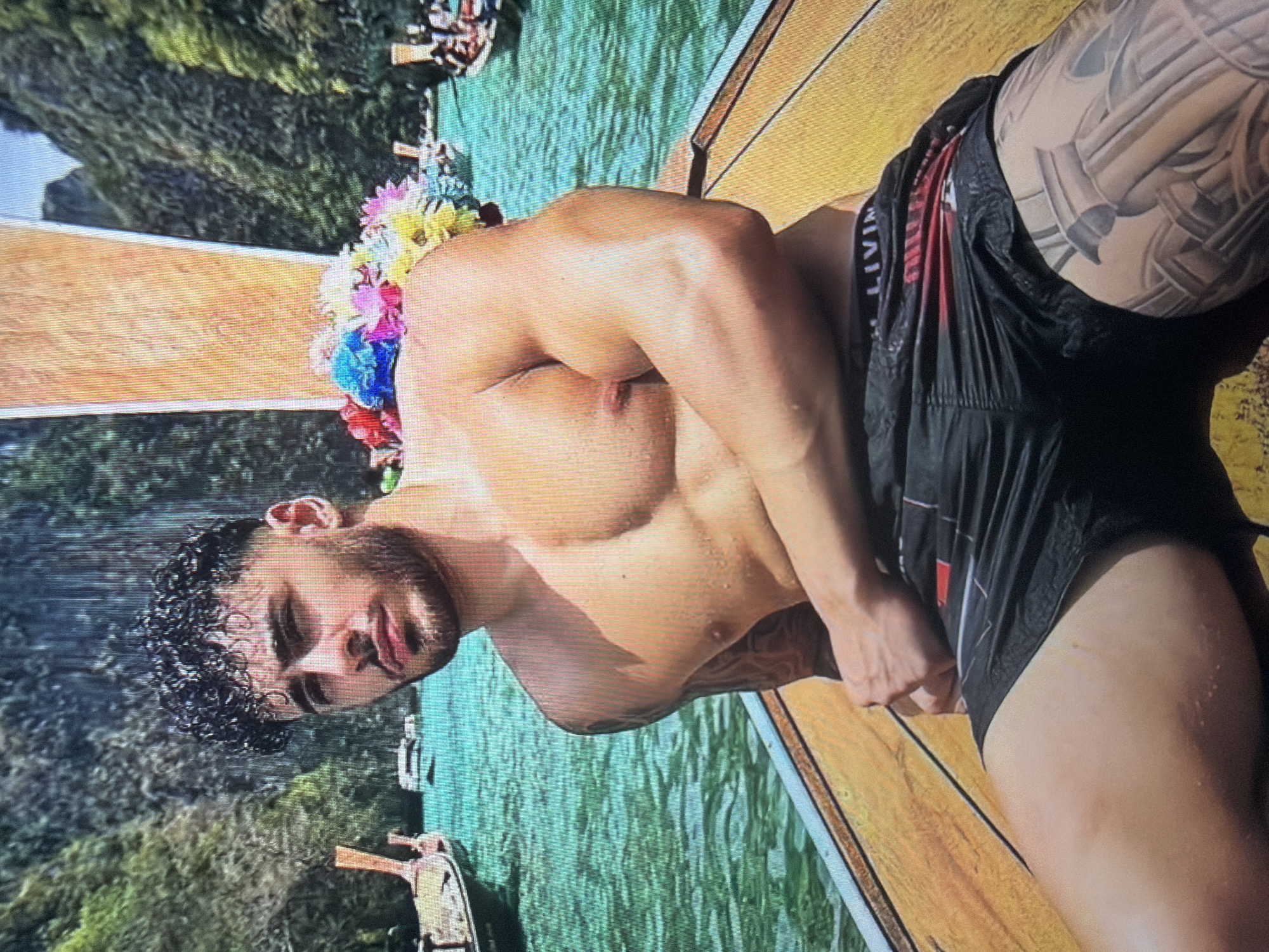 A male individual is depicted sitting at the front of a wooden longtail boat on turquoise water in the city of Shumen, Bulgaria. The man is shirtless, muscular, with dark, curly hair, a beard, and visible tattoos on his left arm and right thigh. He wears black athletic shorts with a red accent and an underwear band showing "LIVIN". He looks directly at the camera with a neutral expression. Colorful artificial flowers are arranged on the boat's bow to his right. In the background, numerous other longtail boats are present on the water, surrounded by steep, verdant, forested limestone cliffs under a bright sky.