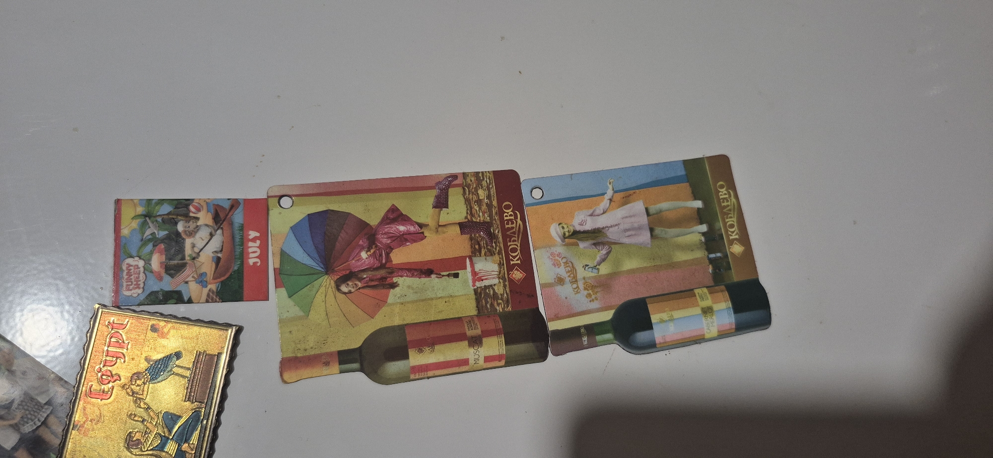 The image shows a flat lay of several novelty items, likely refrigerator magnets, on a white surface. From top left to bottom right, the items are:
1. A golden rectangular magnet with the word "Egypt" in ornate red lettering. The magnet also depicts two figures in ancient Egyptian attire and a bird.
2. A small, rectangular magnet with a cartoon image of a sheep in a rowboat, holding an umbrella, with the text "FUNNY SHEEP" at the top and "JULY" at the bottom.
3. A larger, rectangular magnet featuring a wine bottle on the left and a woman in a pink raincoat holding an umbrella and a paintbrush, with a bucket of paint, on the right. The background has colorful stripes. Text at the bottom reads "КОБАЛЕВО".
4. Another large, rectangular magnet, similar in design to the previous one, also featuring a wine bottle on the left and a young woman in a pink coat and beret holding a can of spray paint on the right. The background also has colorful stripes. Text at the bottom reads "КОБАЛЕВО".
The overall arrangement suggests a collection of decorative items. The white surface is plain and featureless, offering no clues to the specific location. There is no indication of the city of Kryvyi Rih, Ukraine, or any other geographical context in the provided image.