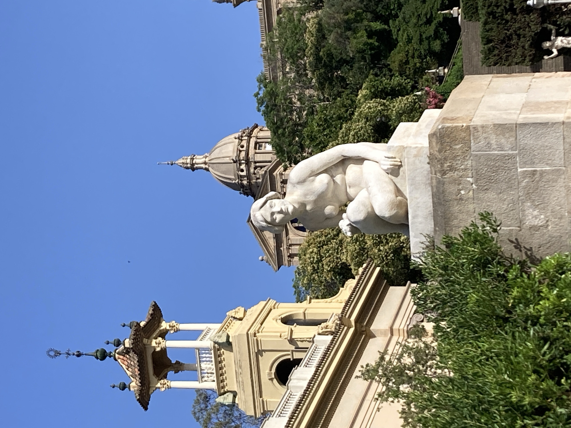 This is a photograph of the Palau Nacional, also known as the National Palace of Catalonia, located in Barcelona, Spain. The building features a large dome, and is situated on Montjuïc hill. In the foreground, a white marble sculpture of a reclining female figure is visible. The sculpture is positioned on a stone pedestal. Behind the sculpture, lush green trees and vegetation are abundant, with the Palau Nacional's architecture visible in the background. The sky is clear and blue, indicating a sunny day. There are no people clearly depicted in the scene.
