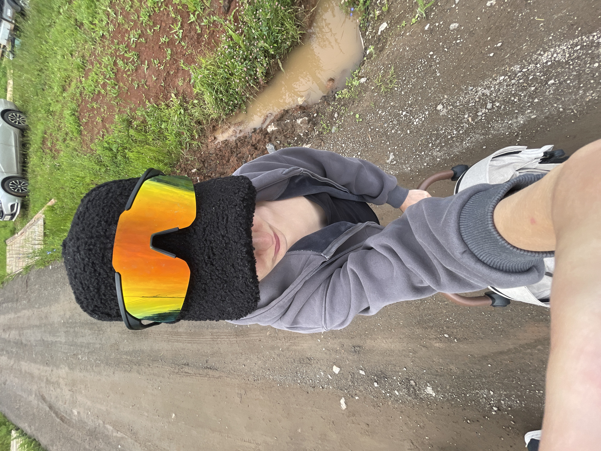 A first-person perspective shot shows a person wearing a black fuzzy hat and reflective orange and yellow sunglasses. They are wearing a grey hooded jacket over a dark tank top. The person appears to be sitting or riding something with a handle, possibly a stroller or bicycle, as part of a light-colored bag is visible below them. The ground is a dirt path with scattered small white stones. To the right of the path is an area of green grass and brown soil, with a muddy ditch containing murky water running alongside the grass. In the background on the left, a silver car is parked on a dirt surface, and on the right, there is more grass and some wooden structures. The overall environment suggests a rural or semi-rural setting, possibly on the outskirts of a village like Podshivalovo, Russia, given the unpaved road and natural surroundings.