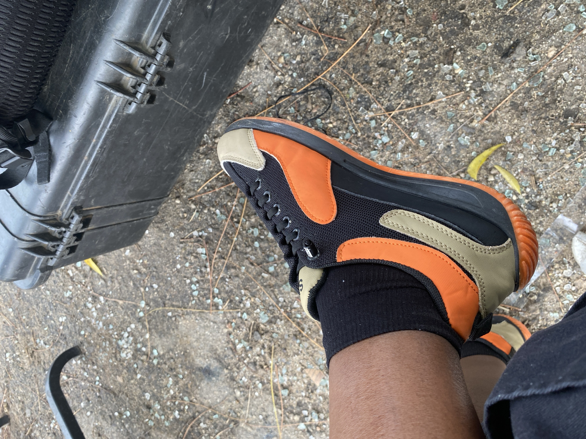 The image shows a person sitting outside, possibly in Lagos, Nigeria, with a black hard case or toolbox next to them. The person is wearing black socks and black pants, with orange, beige, and black sneakers. The ground consists of dirt and small rocks, possibly with broken glass. The environment seems natural, suggesting a possible outdoor or semi-urban area.