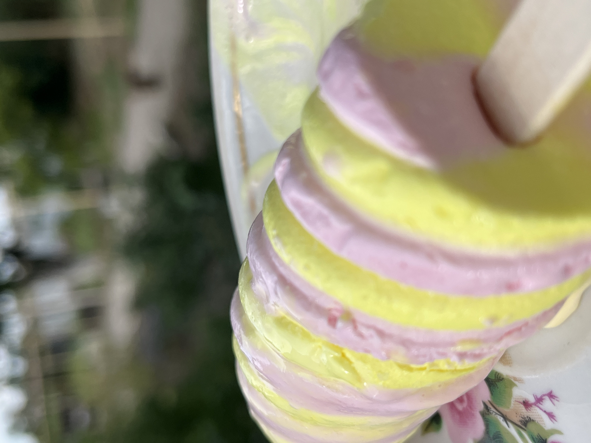 A close-up, slightly blurred shot showcases a swirl ice cream bar with alternating pink and yellow stripes. The ice cream is presented on a white plate decorated with pink flowers. In the background, an out-of-focus outdoor scene depicts greenery and a pathway. No people are visible in the image. The overall lighting suggests daylight.