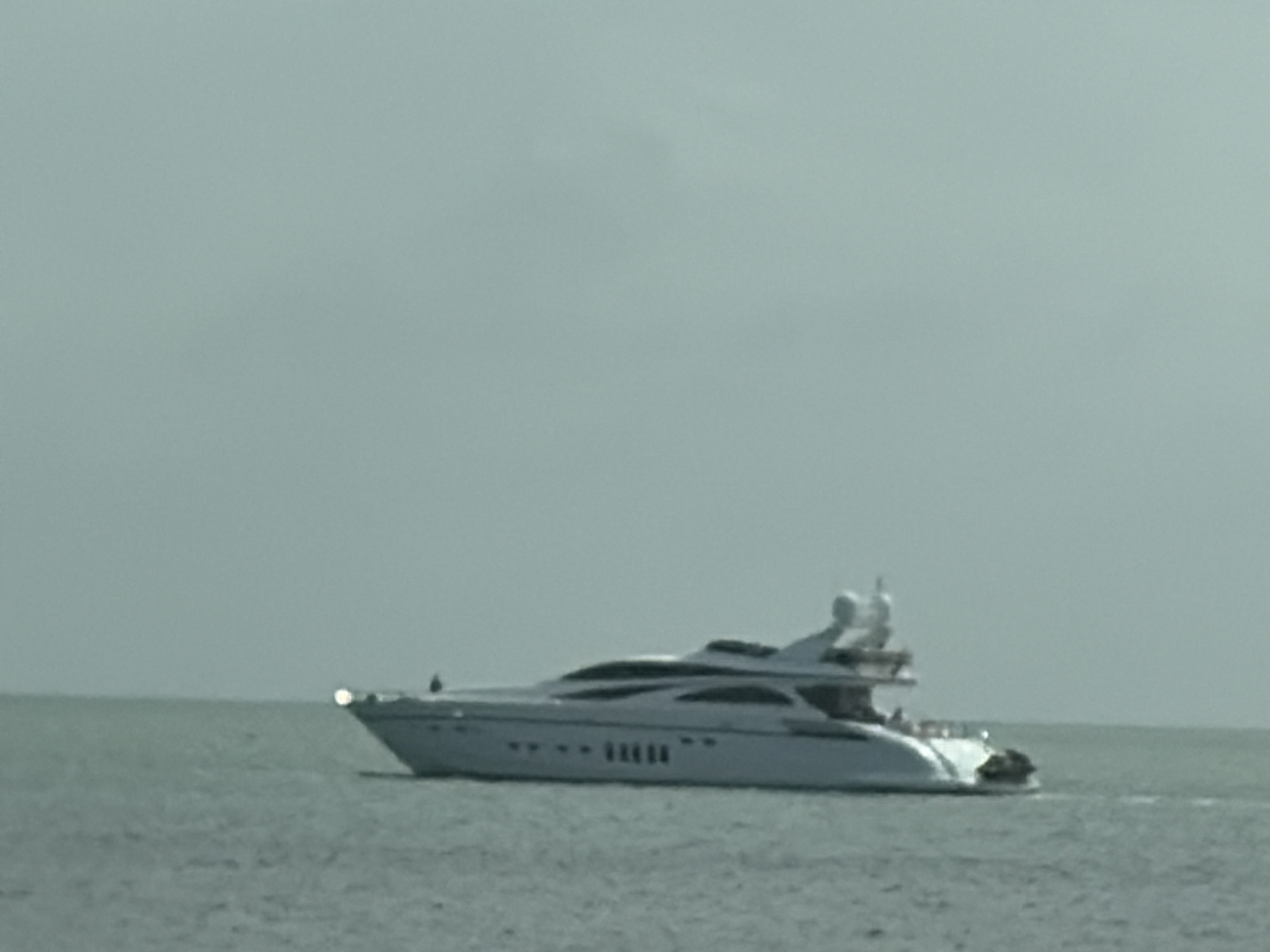 The image shows a white luxury yacht on the water. The yacht is in motion, leaving a wake behind it. The background consists of a light gray sky and a gray body of water. Specific location in Miami is indeterminate based on the image.