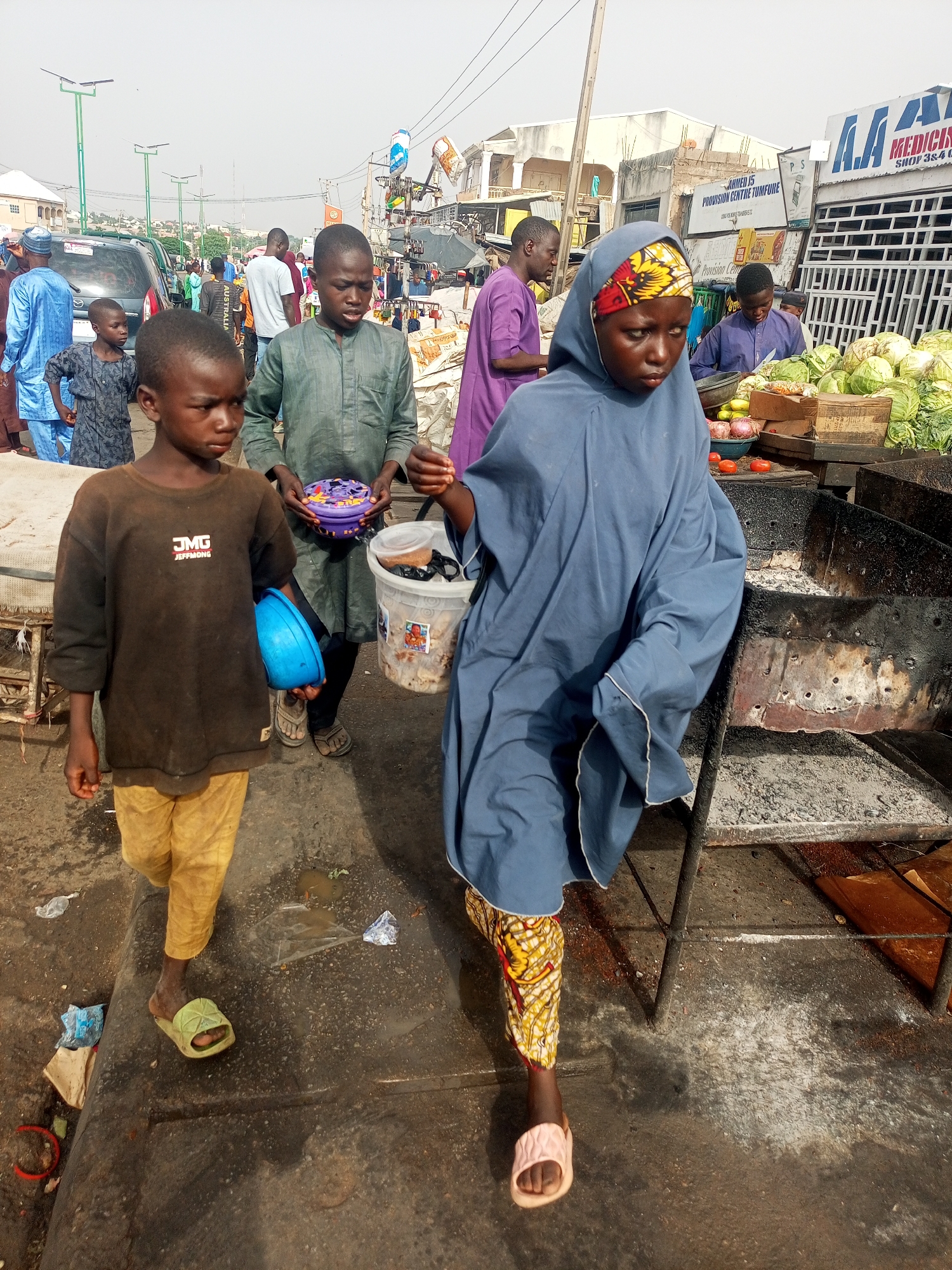 The image depicts three children walking along a street in Tempure, Nigeria. The youngest, a boy, is wearing a brown shirt with "JMG JEFFMONG" printed on it, along with yellow pants and green sandals. He is holding a blue bowl. The second child, a boy, is wearing a green robe and holding a container. The third, a girl, is wearing a blue robe, a head covering, and patterned pants, and pink sandals. They are walking past what appears to be a street vendor selling vegetables; cabbages and tomatoes are visible. In the background, there are buildings, people, cars, and street lights. There's litter on the street. A sign for "Ahmed 15 Provision Centre Tempure" is visible on a building. A sign for "Medicine Shop 3&4" can be seen behind the vendor. The setting suggests a busy market area.