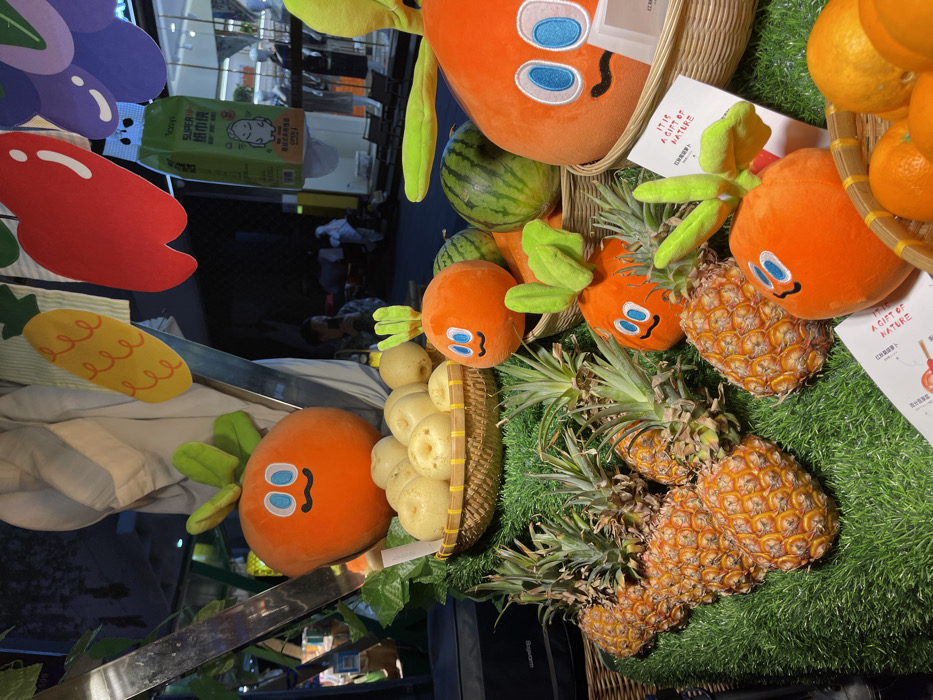 The image is of a fruit stand in Hangzhou, China (30.263, 120.156). The stand is decorated with cartoon orange characters, with the main character being a smiling orange with large blue eyes and a small mouth. The orange character is surrounded by pineapples and watermelons. The fruit stand is situated on a green artificial grass base. In the background, there is a person in a black shirt standing behind a table. The scene takes place in a well-lit, brightly colored environment. The fruit stand is located in a retail setting, possibly within a larger market or shopping mall.