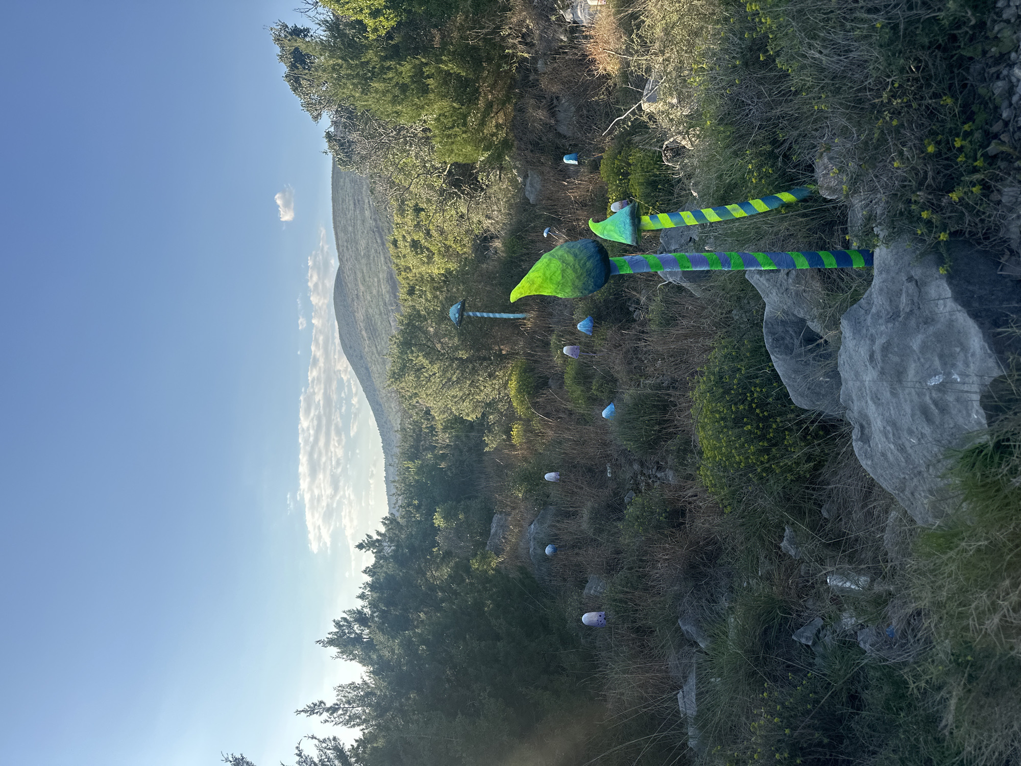The image depicts an outdoor setting in Glavaticici, Montenegro. The landscape features a hillside with vegetation, trees, and rocks. Scattered throughout the scene are colorful, whimsical art installations resembling painted mushrooms or toadstools. A mountain is visible in the background under a blue sky with a few clouds. The art installations are painted in bright colors such as green, blue, and yellow.