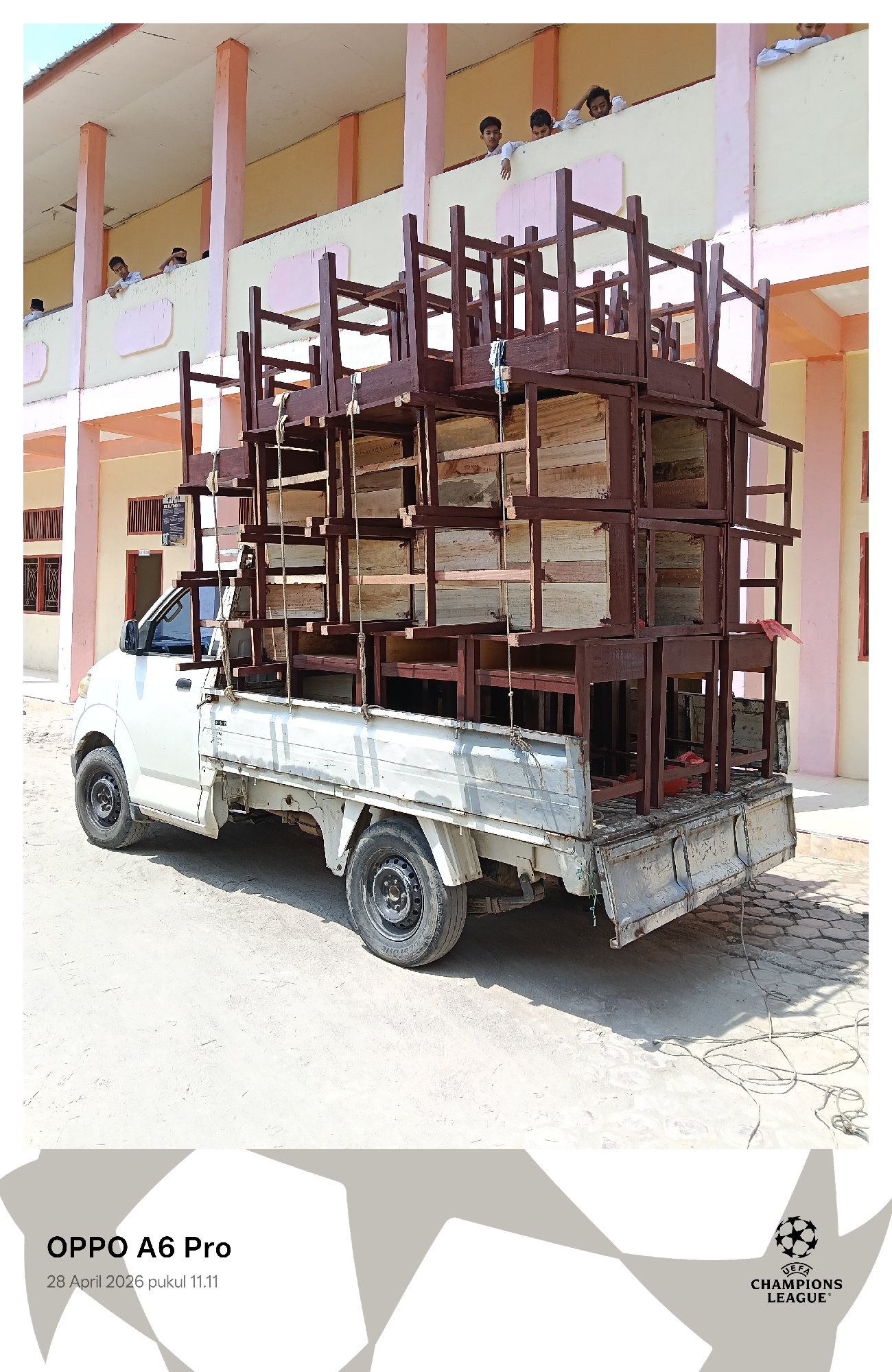 A white pickup truck, heavily laden with numerous dark brown wooden desks and chairs, is parked outdoors on a bright, sunny day. The furniture is stacked high in the truck bed, secured by ropes, with the wooden tabletops of the desks appearing lighter in color.
In the background, a multi-story building with light peach-colored pillars and cream-colored walls serves as the backdrop, suggesting a school or educational institution in Lhokseumawe, Indonesia. Several young boys, likely students in light-colored uniforms, are visible on the second-floor balcony, looking down with curiosity at the loaded truck. The strong sunlight casts clear shadows on the paved ground.
The scene depicts the transportation of school furniture, with the boys observing the activity. Visible text includes "OPPO A6 Pro" and "28 April 2026 pukul 11.11" in the bottom left corner, indicating the camera and timestamp. A "UEFA CHAMPIONS LEAGUE" logo is also present in the bottom right corner.