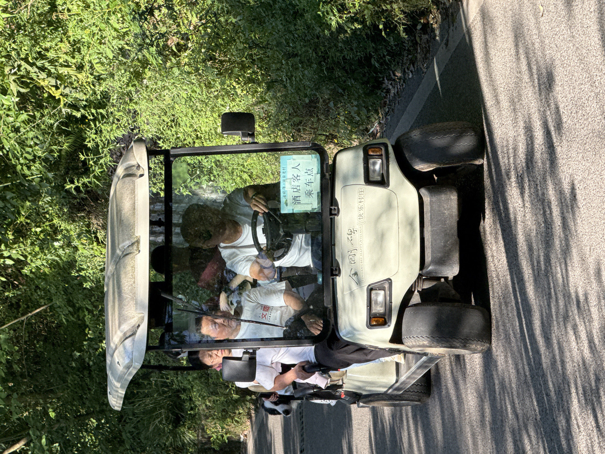 A golf cart with four passengers is traveling on a paved road alongside greenery. The cart has a sign in Chinese indicating it's for hotel guests and a location called "Jingangbei Happy Village", located in Beibei, China. The driver is holding the steering wheel, and the other passengers are seated. The road is casting tree shadows.