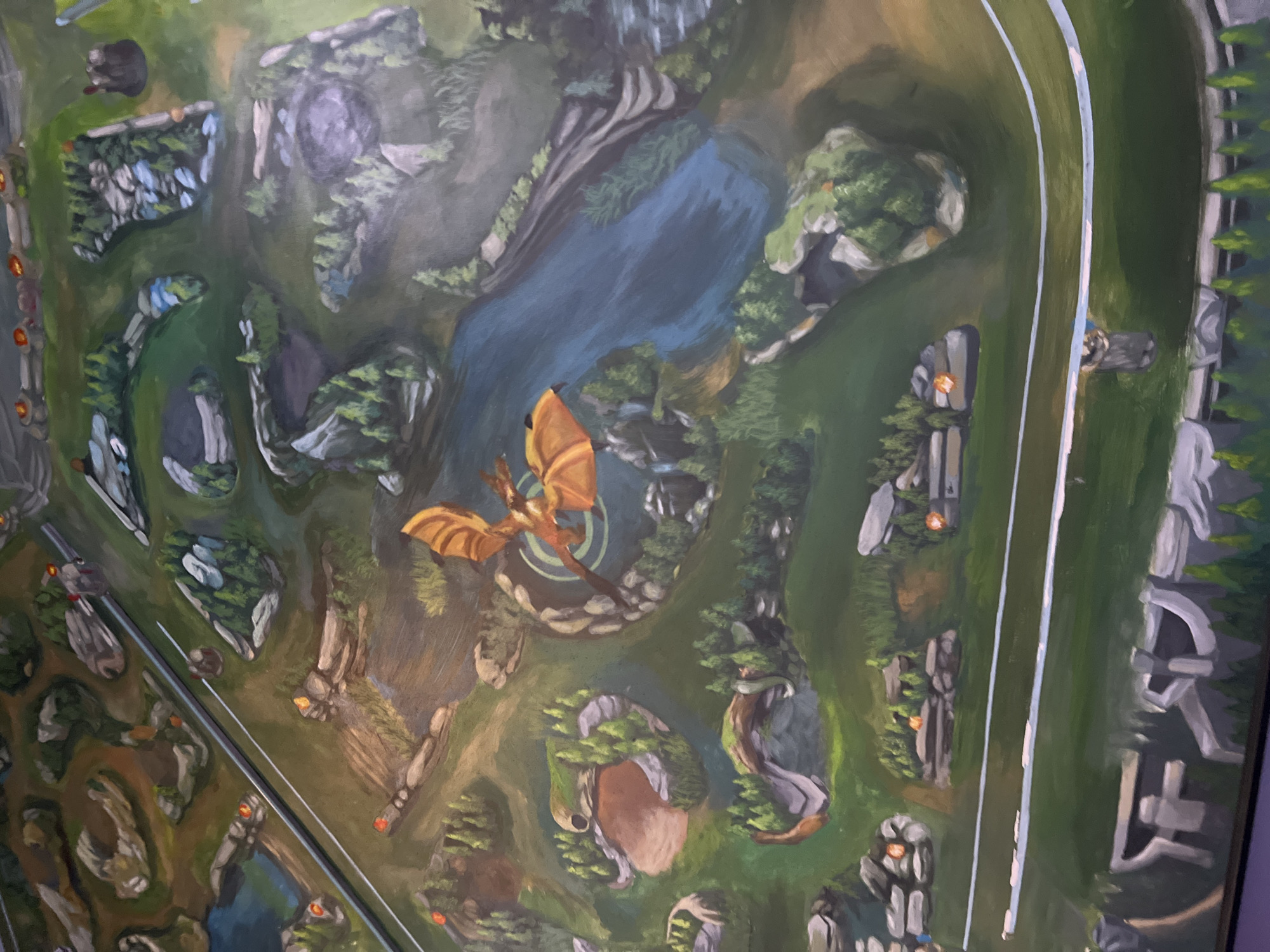 The image is a painting of a fantasy map, possibly a game board, featuring a dragon in flight over a landscape of rivers, rocky outcrops, trees, and what appear to be bases or structures. There are no identifiable landmarks or elements that connect it to the city of Đà Nẵng, Vietnam. The scene is entirely illustrative and does not depict a real-world location.