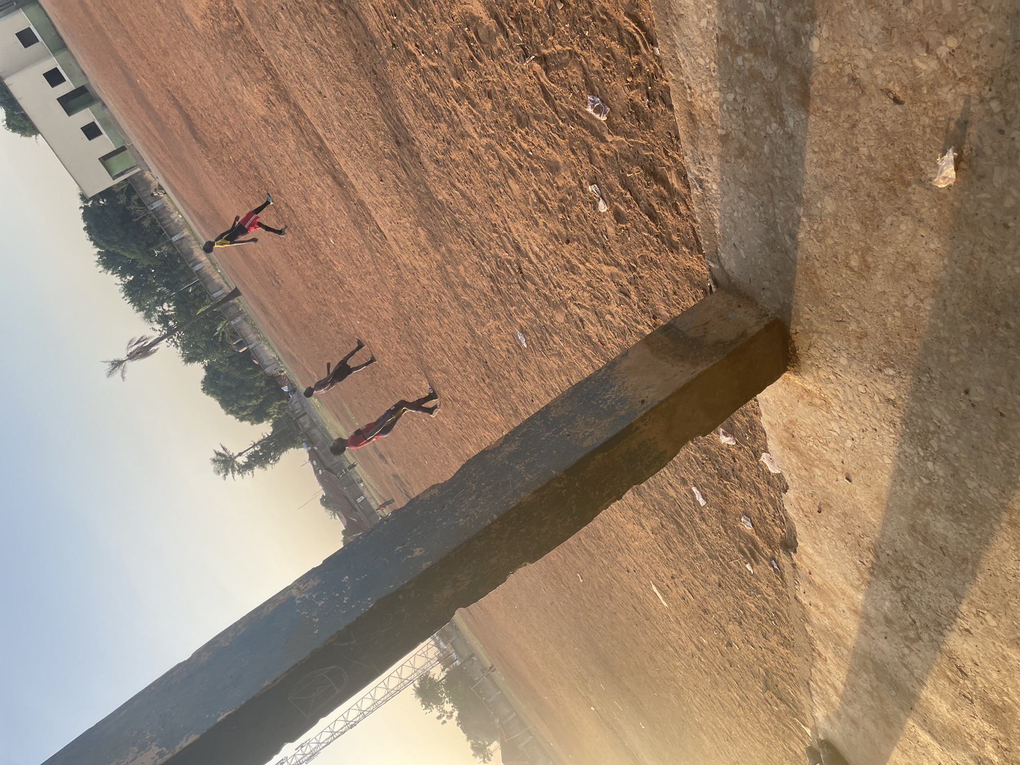 The image captures an outdoor scene, likely a sports field or open ground in Akwanga, Nigeria, bathed in the warm, low-angle light of either sunrise or sunset. A large, dark, and weathered concrete or wooden beam occupies the foreground, extending horizontally across the lower third of the frame. This beam, possibly a barrier or structural element, shows signs of wear and has faint, illegible white markings or a rough sketch on its surface.
Beyond the foreground beam, a vast reddish-brown dirt field stretches into the distance, appearing dry and somewhat uneven. Small pieces of white litter are scattered across the ground. In the middle distance, three silhouetted figures, appearing to be young people or children, are actively moving across the field from left to right. One individual is visible in a yellow top and red shorts, another in a dark outfit, and a third in a red top and dark shorts, all appearing to be running or playing.
The background features lush green trees, including distinct palm trees. A light-colored building with several dark, square-shaped windows or openings is visible on the far left. The sky transitions from a clear light blue at the top to a golden, yellowish-orange hue near the horizon, indicative of the time of day. A long shadow cast by the foreground beam extends onto the adjacent ground, further emphasizing the early morning or late afternoon sun. A partial metal structure, possibly a fence or part of an overhead utility, is visible near the horizon on the right. The overall impression is an active, open-air environment during a peaceful time of day.