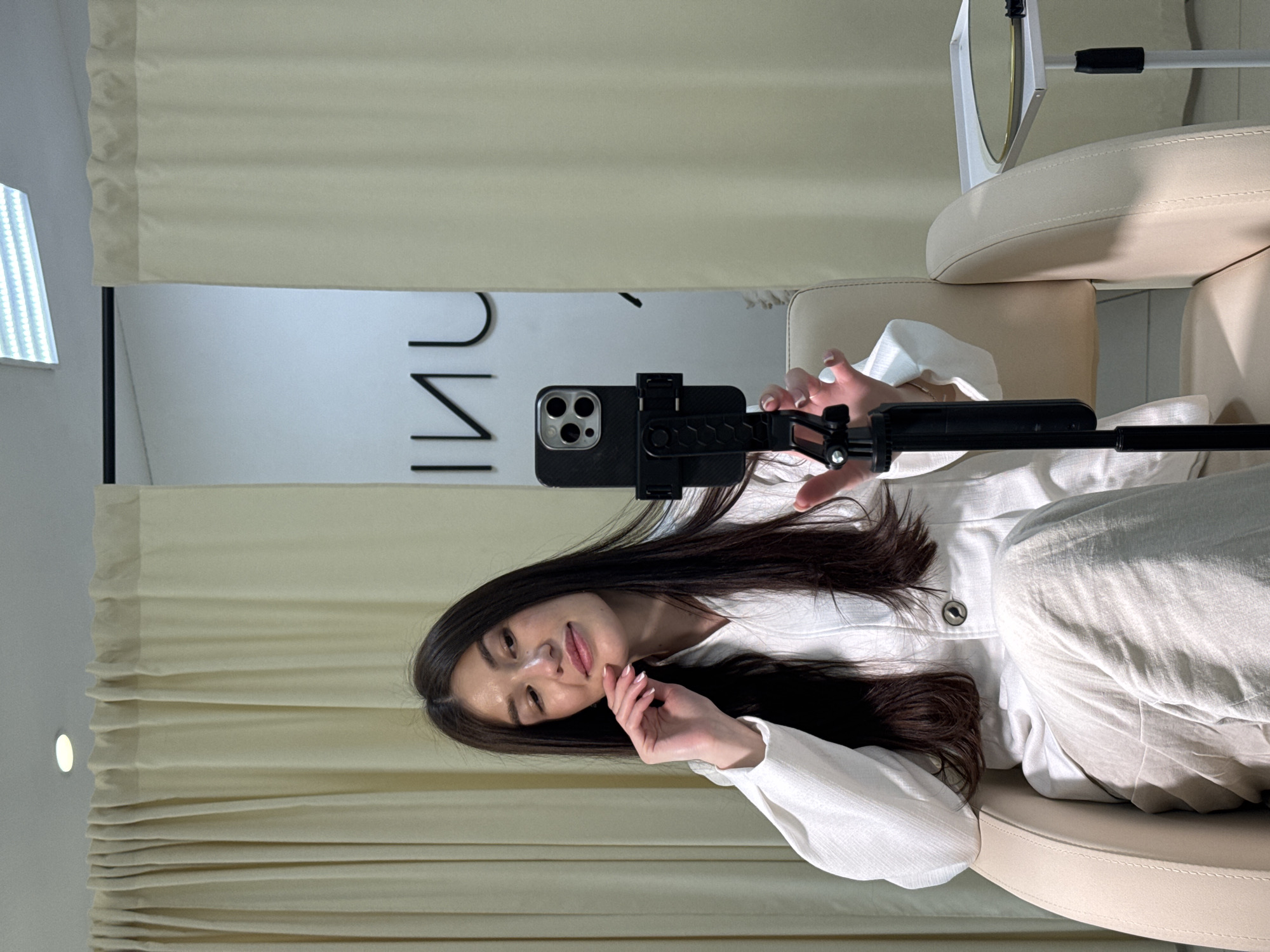 The image, oriented horizontally, depicts a young woman in an indoor setting, appearing to be sitting upright in a light-colored armchair if viewed correctly. She has long, dark hair and is wearing a white long-sleeved top and light-colored pants. The woman is holding a smartphone, likely an iPhone, attached to a black selfie stick or tripod, directing it towards herself. Her left hand is gently positioned near her chin, and she looks towards the camera with a soft, pleasant smile.
The background features light beige blinds or curtains on the left (if upright). Behind the woman, on a white wall, large, dark, sans-serif letters are visible, spelling out "ИНГ" (ING), suggesting a business name or sign. Further to the left, a bright LED light panel is partially visible. To the right of the woman, part of another light-colored armchair and a white-framed mirror on a stand can be seen. The scene conveys a clean, modern aesthetic, possibly a studio, salon, or clinic. Given the location context of Chaikovsky, Russia, the Cyrillic text is appropriate. The overall mood is calm and inviting.