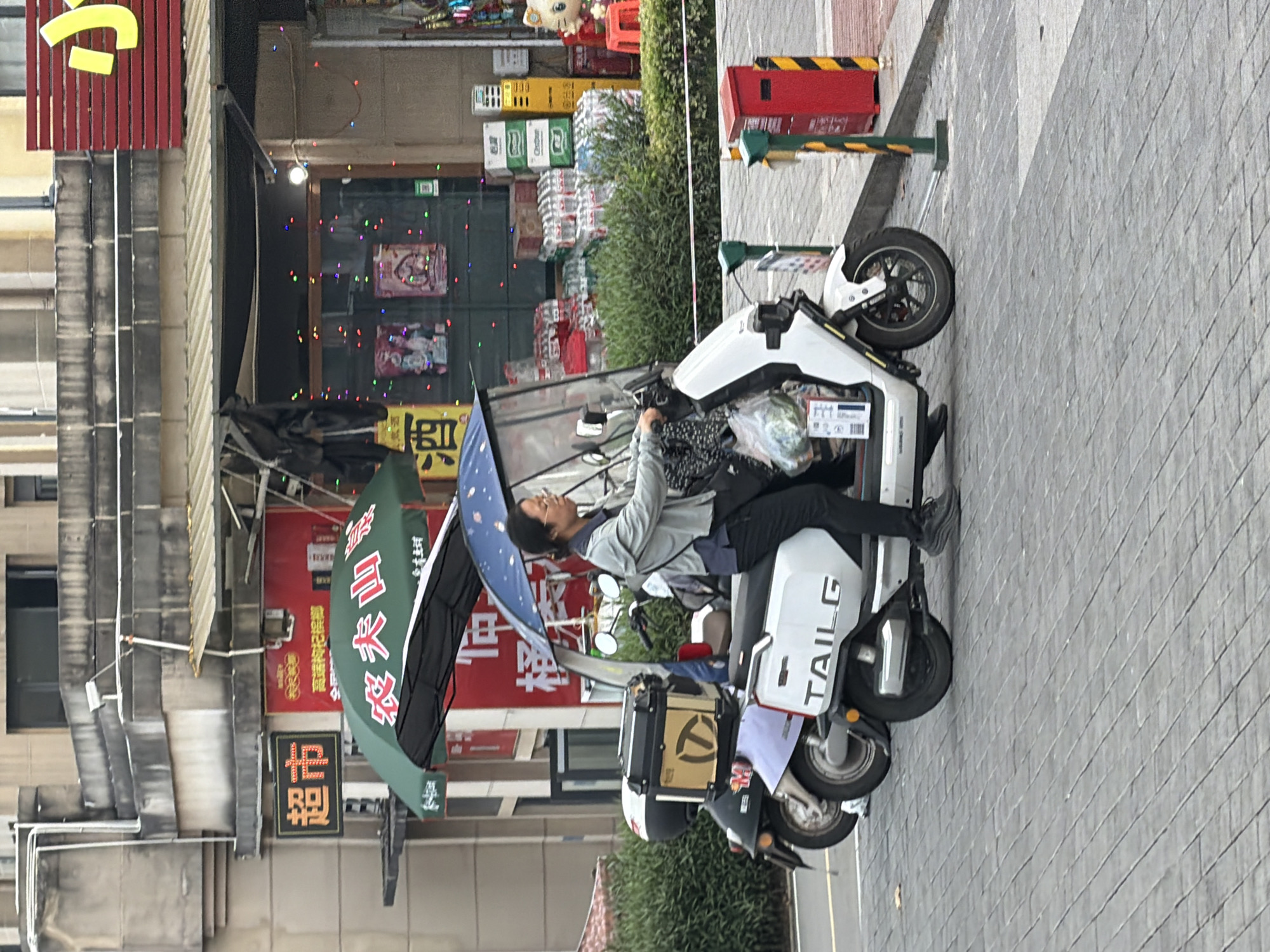 A person is riding a white electric scooter on a paved city street in Jiangjin, China. The scooter has a large green umbrella attached for shade, and a clear windshield. The rider is wearing glasses, a grey jacket, and black pants. They are holding a plastic bag of groceries. Another scooter, partially visible, is parked to the left. In the background, there are buildings with storefronts, including one with Chinese characters that read "超市" (supermarket) and another with a red sign featuring yellow characters. There are stacks of bottled drinks and boxes outside one of the stores, decorated with colorful lights. A red trash can is visible on the sidewalk to the right. The street is paved with grey interlocking bricks.