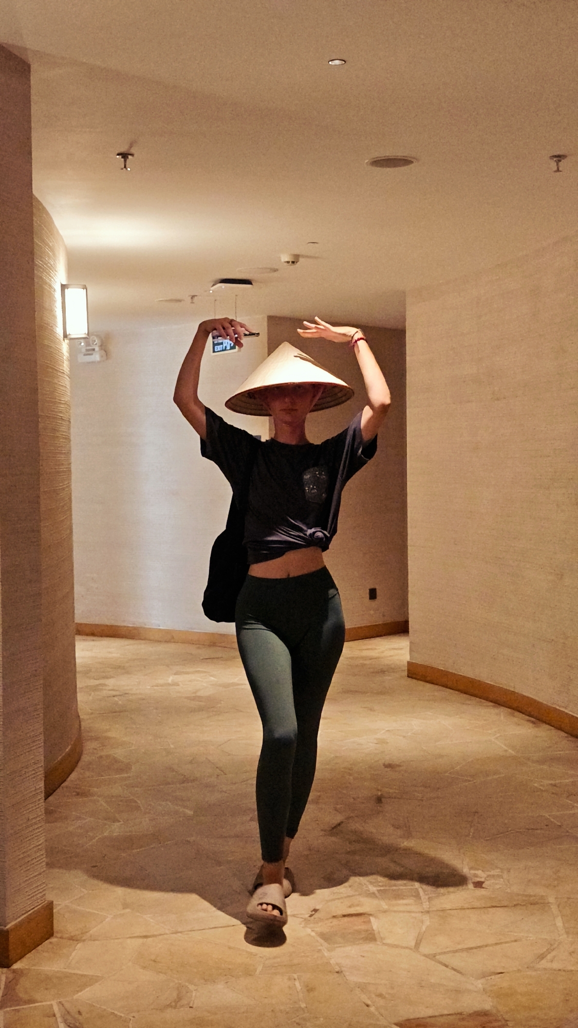 A person, likely female, stands centrally in a warmly lit, curved hallway, striking a dynamic pose. They wear a dark t-shirt, knotted at the waist to expose their midriff, dark green leggings, and light-colored slip-on sandals. A traditional Vietnamese conical hat (Nón Lá) covers their head, obscuring their face. Both arms are raised above their head; the left hand holds a smartphone with the word "EXIT" clearly visible on its screen, while the right hand is posed gracefully with a red bracelet on the wrist. A dark bag or backpack is slung over their left shoulder.
The hallway features light-brown, textured walls and a floor made of irregularly shaped, light-colored tiles. Recessed ceiling lights and a wall-mounted light fixture illuminate the space, casting soft shadows. In the distance, a blue/green "EXIT" sign is visible. The architectural style suggests an indoor setting like a hotel or resort in Mu Cang Chai, Vietnam. The person's pose, combined with the traditional hat, evokes a sense of playfulness or artistic expression within this modern interior. The warm artificial lighting suggests it could be evening or night.