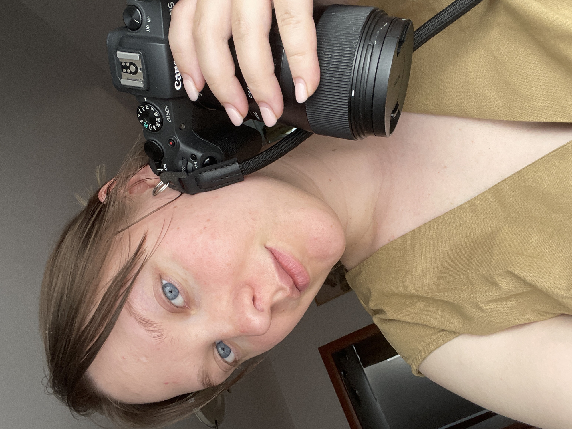 The media file contains a selfie taken by a person holding a black Canon EOS RP camera with a lens attached. The person has blue eyes and light brown hair, and is wearing a tan-colored top. The camera is positioned in the foreground, partially obscuring the person's face and upper body. The background is blurry and appears to be an indoor setting with a plain wall. Visible text on the camera includes "Canon," "EOS RP," "Av," "M," "SCN," and "SIGMA." The lighting suggests it is daytime, possibly indoors. The person's expression is neutral. The overall impression is that of a casual self-portrait taken by a photography enthusiast.
