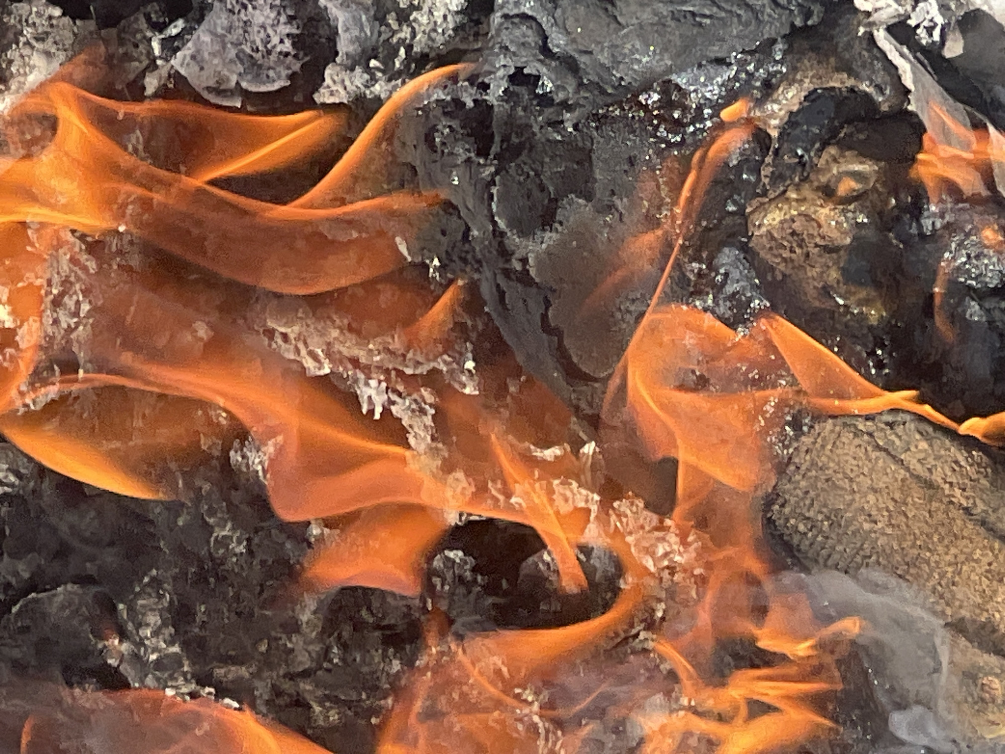 The image depicts a close-up of a fire burning amidst charred wood and ash. Flames are prominent, appearing orange in color. Small amounts of ash are visible. The location in the city of Gura Top, Nigeria is not discernible from the image.