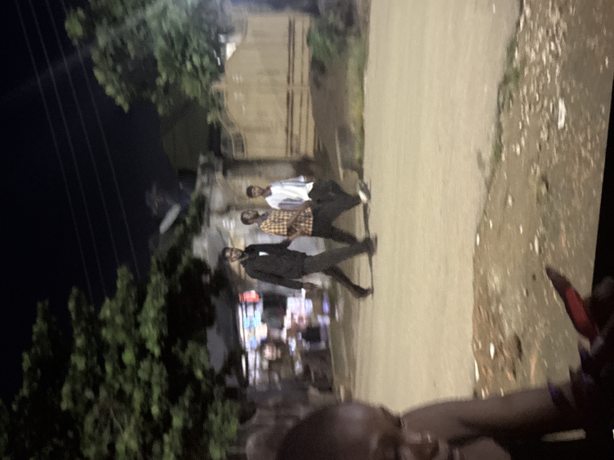 The image depicts a nighttime street scene in Abuja, Nigeria. Three men are walking on a paved road. The man on the left wears a black hooded jacket and dark pants. The middle man is wearing a plaid shirt and dark pants. The man on the right is wearing a light-colored long-sleeved shirt and dark pants. In the background, there are trees, buildings, and a fence. The road appears to be dusty and uneven, with some debris scattered along the edges. The lighting suggests artificial illumination from streetlights or nearby establishments. A portion of what appears to be a person's head and shoulder, possibly a woman, is visible in the foreground on the left side of the image, out of focus.